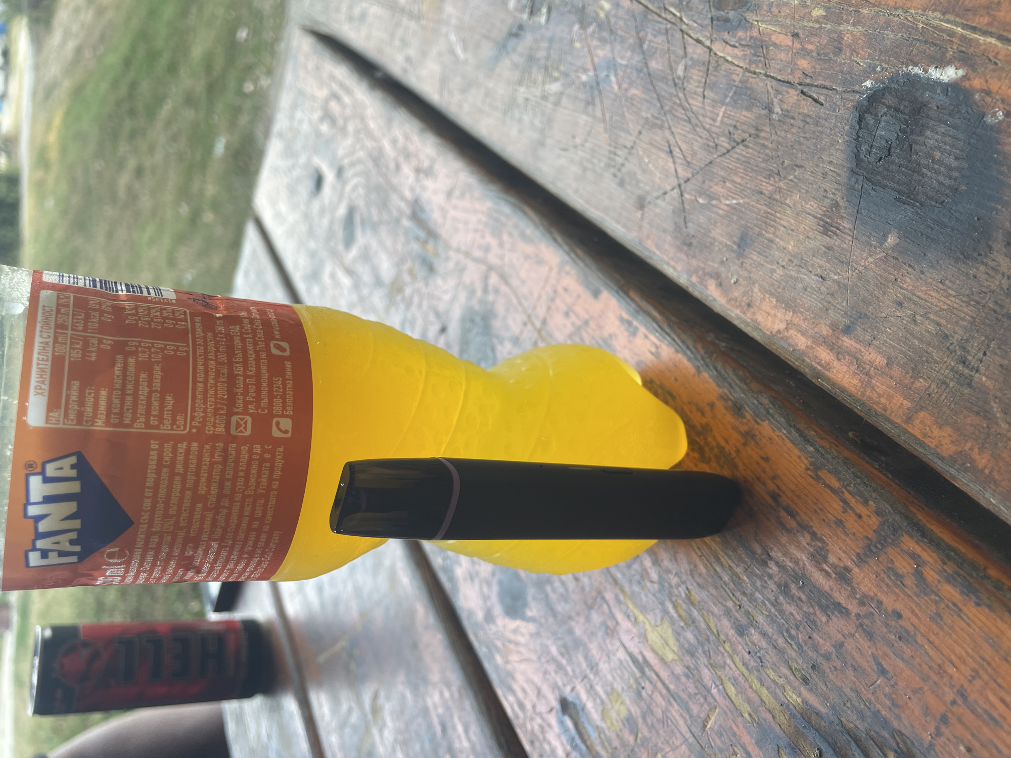 The image depicts an outdoor scene on a wooden table. A dark-colored electronic cigarette device stands upright in the foreground. To its right, a partially visible Fanta bottle, filled with a yellow liquid, is placed on the table. Behind the Fanta bottle, a red and black can, likely a beverage, is also present. The table appears weathered, with visible grain and some scratches. In the background, greenery and a blurred outdoor setting are discernible, suggesting a park or recreational area. No people are clearly visible in the frame, though a blurred arm can be seen on the far left. The location is specified as Blagoevo, Bulgaria.