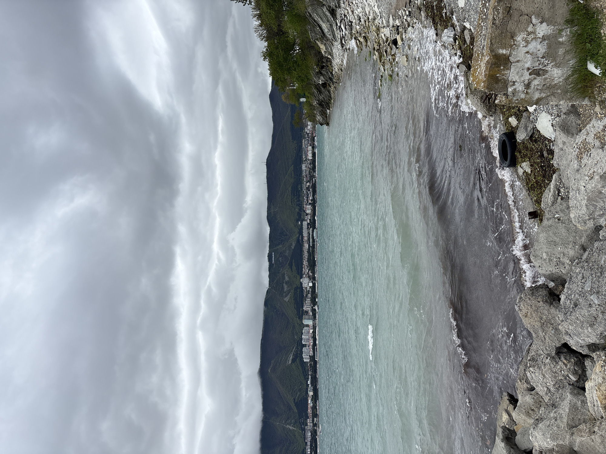 The image depicts a cloudy coastal scene in Gelendzhik, Russia. A city skyline is visible at the base of a mountain range that stretches along the coast. The sky is filled with dense, striated clouds. The sea extends to the right, with varying shades of grey and turquoise. Along the shoreline, a rocky embankment is visible. A tire is situated amongst the rocks near the water's edge. The embankment consists of a mix of grey rocks, concrete, and some vegetation. The image shows no people, but the built environment and coastline are clearly depicted.