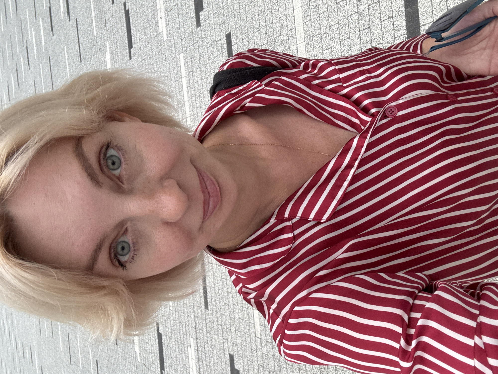 A close-up, overhead selfie of a light-skinned Caucasian woman with blonde bobbed hair and green eyes. She is wearing a red and white striped button-down shirt and a delicate gold necklace. The background appears to be an outdoor tiled walkway with a repeating pattern of light and dark rectangles. The woman is smiling subtly and holding a pair of light blue sunglasses in her right hand, partially visible in the lower right corner of the frame. The overall setting suggests an urban environment.