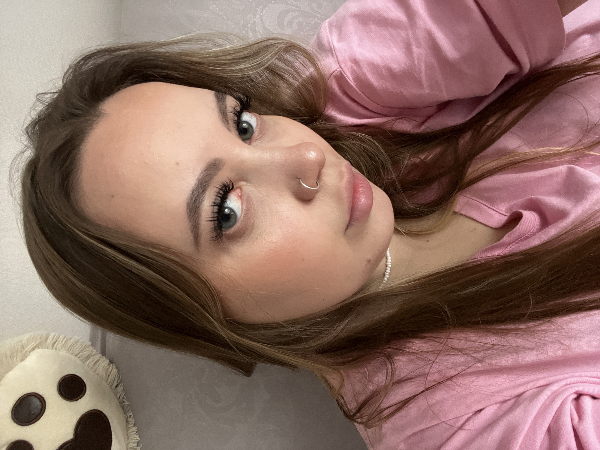 A close-up, horizontal portrait features a young woman lying down, looking directly at the camera. She has long, wavy brown hair with lighter highlights, which is spread around her and partially rests on a pink t-shirt. Her eyes are a striking green-blue, emphasized by long, dark eyelashes and well-defined eyebrows. A small silver hoop nose ring adorns her left nostril. Her skin appears smooth, with a subtle blush on her cheeks and full, slightly glossy lips. She wears a simple white beaded necklace. The woman is dressed in a plain pink t-shirt. In the bottom left corner, a portion of a white pillow with dark brown paw print designs is visible. The background is a light-colored, possibly white, wall with a faint decorative texture. The lighting suggests an indoor setting. The woman's expression is calm and neutral. No text is visible. The location context provided is Krasnodar, Russia.