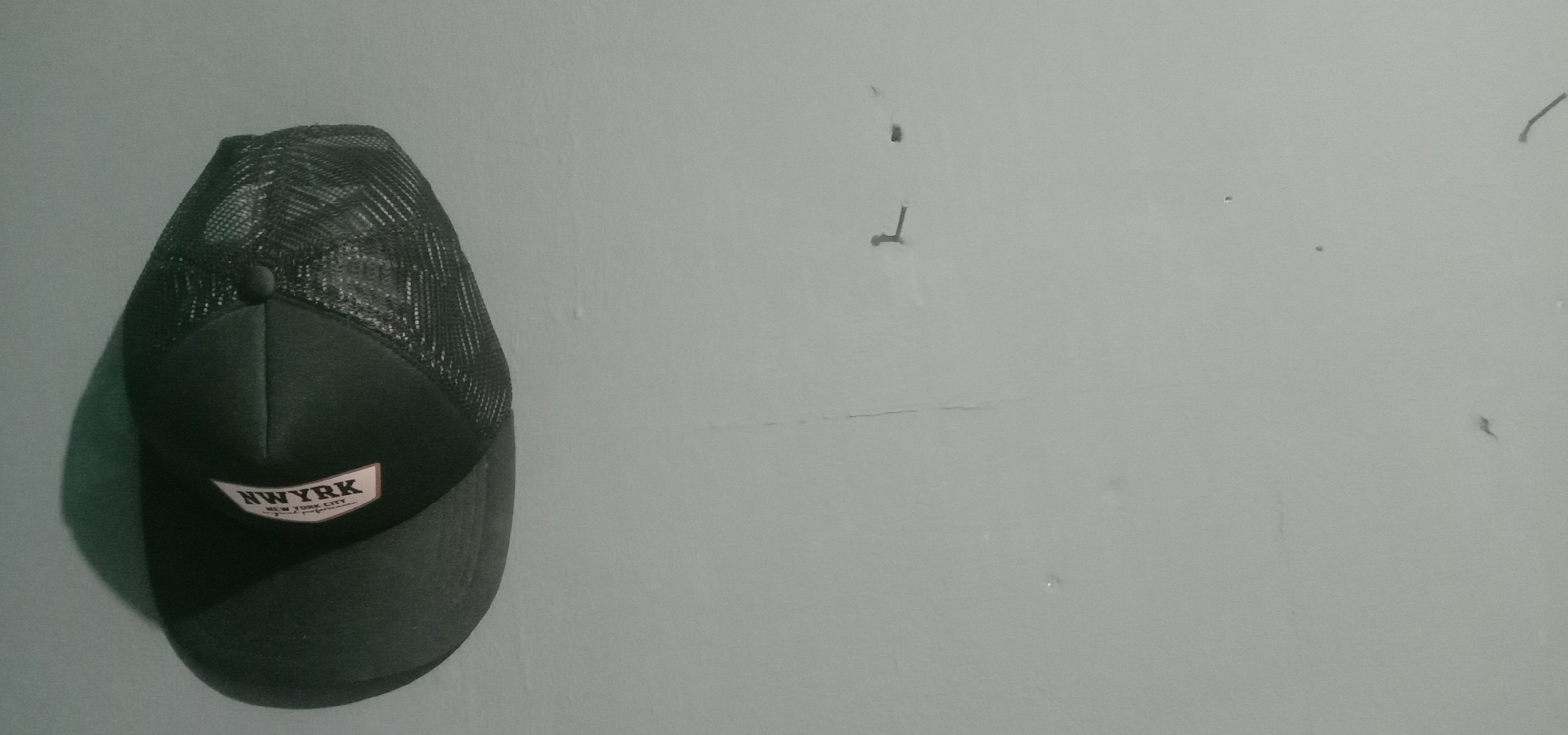 A dark colored baseball cap with a mesh back is positioned against a light gray wall. The cap has a label with text that reads "NWYRK NEW YORK CITY" in white and black. The wall appears to be indoors, and has several small marks and one visible crack. There are no people present in the image. The location of the image is unknown, and there is no information to suggest it is in Kendari, Indonesia.