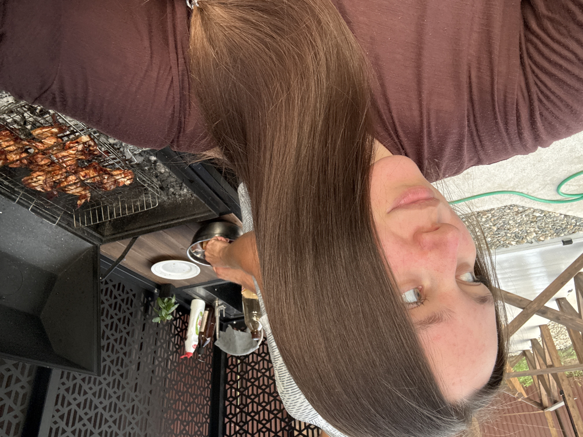The image shows a young woman with long brown hair, wearing a brown t-shirt, taking a selfie outdoors. She is partially obscured by her hair. In the background, there is a black grill with chicken cooking on a rack. There are also some bottles and a plant on a counter next to a sink. A wooden fence and some gravel are visible in the foreground. The location is identified as Starokorsunskaya, Russia.