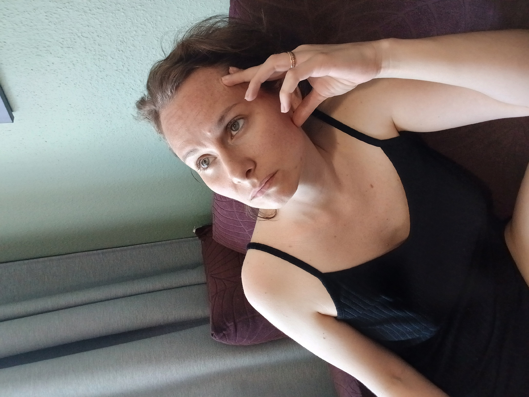 A woman with brown hair and light skin is looking upwards with a thoughtful expression, resting her forehead with her right hand. She is wearing a black tank top and a gold ring on her right hand. The woman is seated on a purple couch, with dark green curtains and a light green wall visible in the background. The image appears to be taken indoors in a domestic setting. There are no identifiable landmarks or elements to suggest a specific location within Yekaterinburg, Russia.