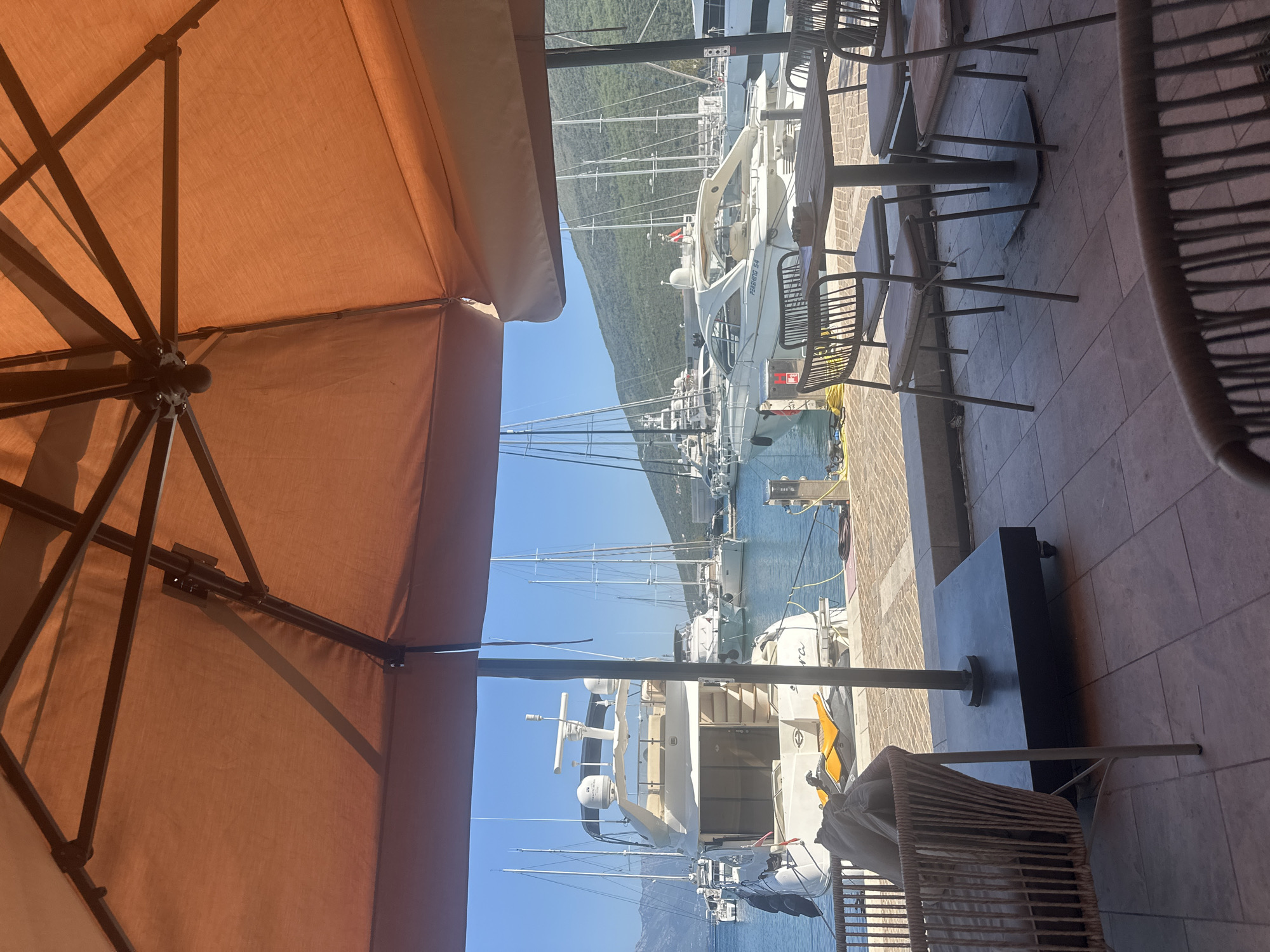 This image captures a view of a marina from a shaded outdoor seating area. The foreground features an orange umbrella and outdoor furniture, including a woven armchair with cushions and a table with chairs. Beyond the seating area, the marina is filled with various yachts and sailboats docked in calm blue water under a clear sky. Green, tree-covered hills form the backdrop of the scene, suggesting a coastal or waterfront location. This view is consistent with the appearance of the waterfront promenade in Herceg Novi, Montenegro, known for its numerous yachts and scenic surroundings.