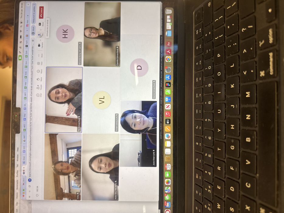 The image displays a dark-colored laptop, consistent with an Apple MacBook, actively running a Microsoft Teams video conference. The laptop's keyboard, featuring a UK layout with a '£' symbol, is prominent in the foreground. The screen's MacOS menu bar shows the date and time as "Thu 29 Jan 09:08" and a series of application icons in the dock below the main display area. A partial reflection of a person's head is visible on the upper bezel of the laptop screen.
The video conference window presents seven participants in a grid. On the top row, from left to right: Kitty, a female with dark hair wearing a light hooded top, is shown against a background of wooden beams and a window; Flo (Unverified), a female with dark hair and a grey sweater, is actively gesturing with her right hand against a plain light wall; and a placeholder circle with the initials 'HK'.
The middle row features: Kobo Siu, YN (MTI), an East Asian female with dark hair in a dark top, depicted with her mouth slightly open against a plain light wall; a placeholder circle with the initials 'VL'; and Camilla (Unverified), a female with long dark hair, wearing glasses and a dark top, against a subtly blurred light background.
The bottom row displays: June Chan (LN), an East Asian female with short dark hair, wearing a headset with a microphone and smiling, against a background featuring a light wall with a dark blue element; and a placeholder circle with the initial 'D'.
The Microsoft Teams interface controls for chat, people, reactions, view, camera, microphone (indicated as muted), share, and leave are visible at the top of the conference window. The meeting duration is displayed as "00:34". Several browser tabs are open above the Teams application, including multiple instances referencing "teams.microsoft.com", "HKT", and "EYT".
The scene is situated in the City of London, United Kingdom.