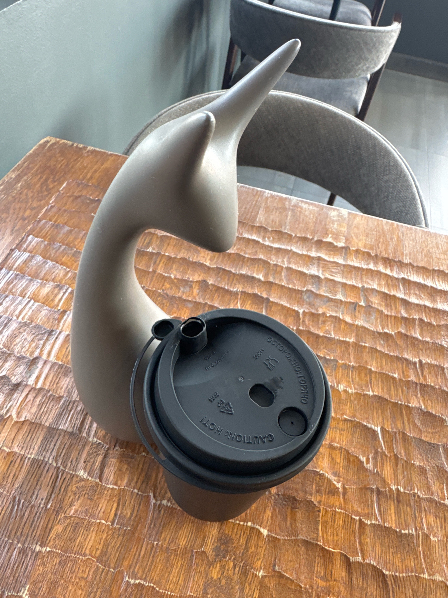 The image depicts a still life composition on a textured wooden table. A grey, abstract sculpture, resembling a stylized bird or dolphin with a long neck and pointed head, is positioned on the left side of the frame. A black disposable coffee cup lid with various inscriptions is placed on the table in front of the sculpture, obscuring the upper part of the coffee cup itself. The lid has two openings, one for drinking and a smaller one for ventilation. In the background, out-of-focus, are upholstered chairs, suggesting an indoor seating area, possibly a cafe or restaurant. The lighting is soft and natural, indicating an ambient indoor setting. The overall impression is one of quiet repose, with everyday objects arranged in an aesthetically pleasing manner. The scene does not provide any specific clues about its location in Moscow, Russia.