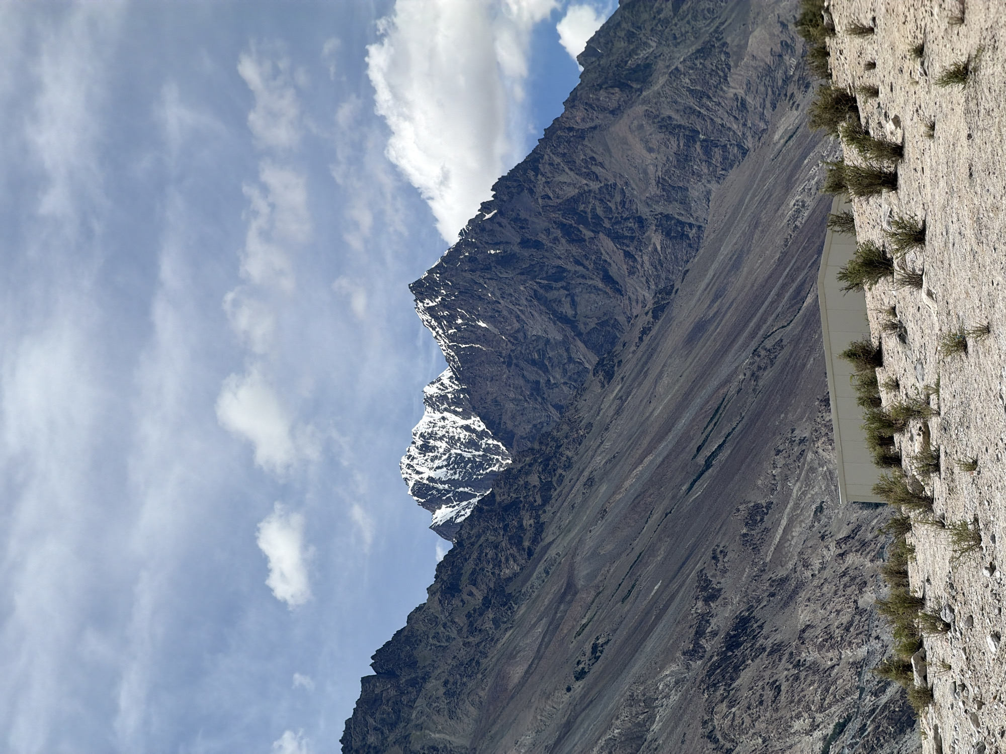 The image shows a wide shot of a mountainous landscape in Panamik, India. The foreground is a barren, rocky terrain with sparse, dry vegetation. A light-colored, rectangular building with a slightly curved roof is situated in the middle ground, partially obscured by the vegetation. In the background, a massive mountain range dominates the scene. The peaks of the mountains are rugged and dark, with patches of snow clinging to the upper slopes. The sky is partly cloudy with scattered white clouds against a pale blue backdrop. The overall impression is one of a remote and arid environment.