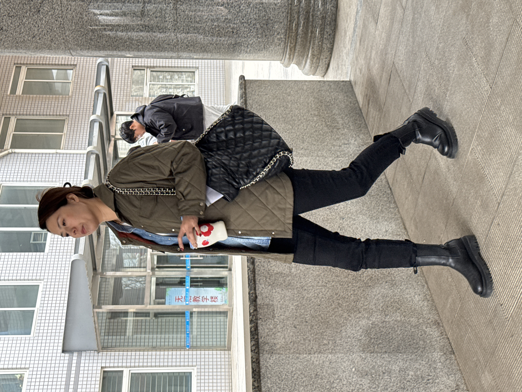 A medium shot presents a woman walking near a building in Zhongtun, China. The woman wears black jeans, black boots, and an olive jacket. She carries a black purse and a white object with red accents in her right hand. Behind her, another individual is visible, wearing a black jacket. The environment consists of a tiled building facade with windows, a gray sidewalk, and stone pillars supporting an overhang. A blue sign with Chinese characters is affixed to one of the building's windows.