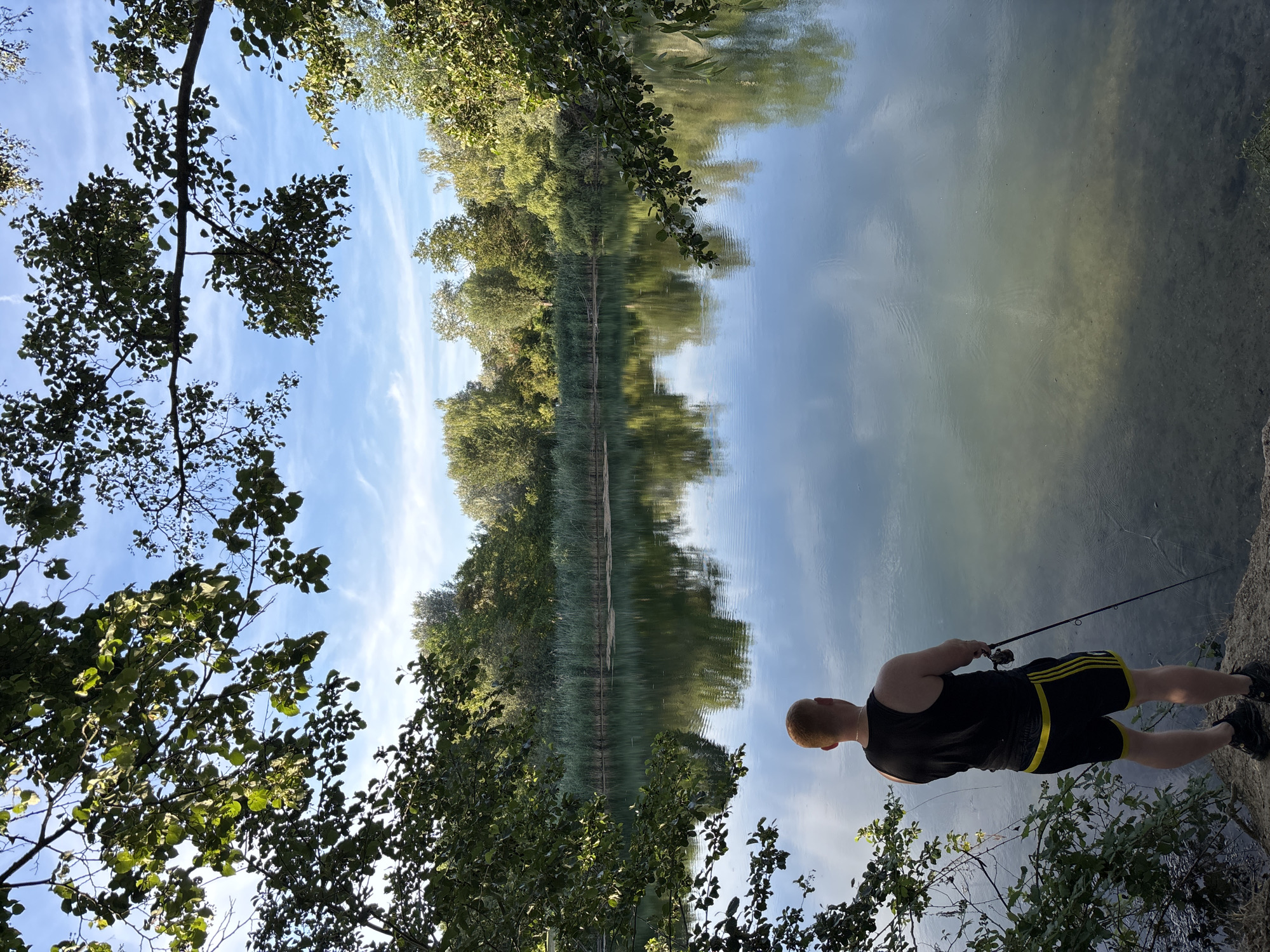 A man with red hair is standing on the bank of a lake in a park, fishing. He is wearing a black tank top and black shorts with yellow stripes. The lake is surrounded by trees and reeds. The sky is blue with some clouds. The scene is peaceful and serene. This lake is located in the city of Ludwigshafen am Rhein, Germany.