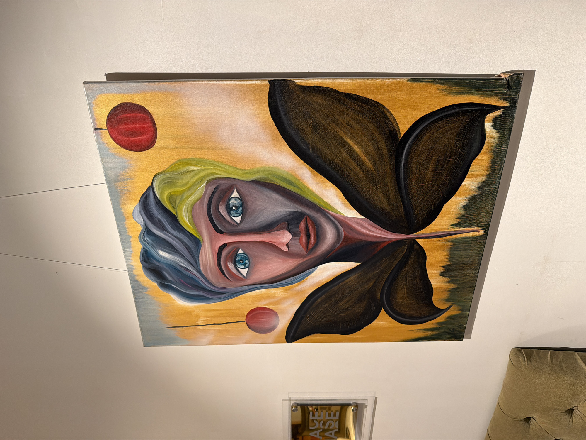 The image shows a framed, rectangular painting mounted on a white wall. The painting is rotated such that the bottom edge faces the right of the frame. The painting depicts an abstract portrait of a face with large, stylized, black wings emanating from the head. The face is pale pink and grey, with blue eyes, red lips, and an elongated nose. The hair is styled in waves, with portions colored green and blue-grey. Two red circles are located on either side of the face against a background of muted yellow and brown hues.
The painting is mounted on a white wall and is positioned above a translucent, rectangular object with gold-colored objects within. A tufted, tan-colored piece of furniture is seen on the right. There is no evidence to indicate this image was taken in the city of Moscow, Russia.