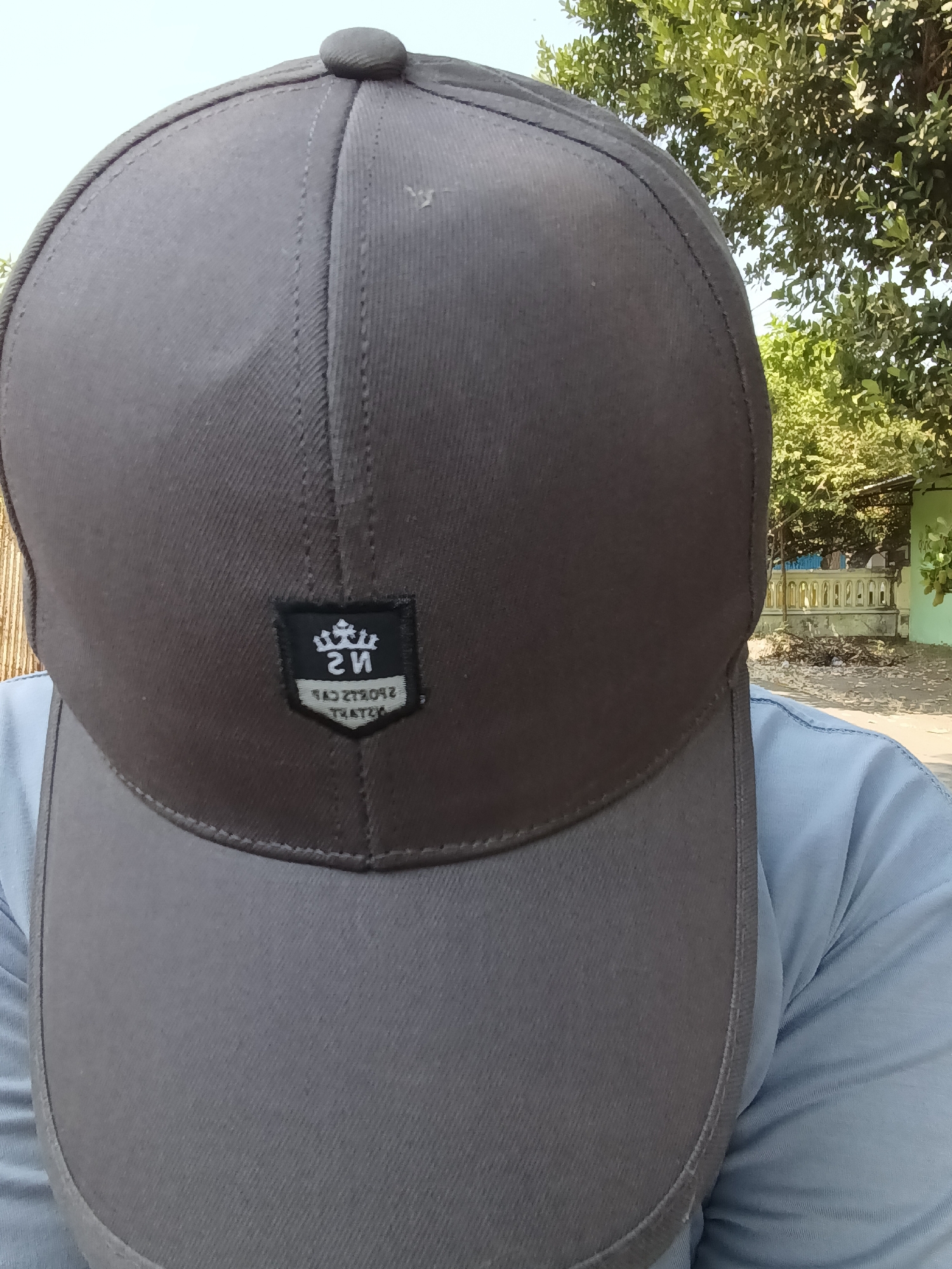 A close-up selfie shows a person wearing a dark grey baseball cap that obscures their face. A small black patch with a white border is affixed to the front of the cap, featuring a crown-like symbol and reversed text. The person is wearing a light blue t-shirt. The background consists of foliage from trees and a pale blue sky, suggesting an outdoor setting during daylight. In the distance, a low-lying wall with vertical posts is visible, along with a light green wall and some brown debris on the ground. The image appears to have been taken from eye level, looking upwards.