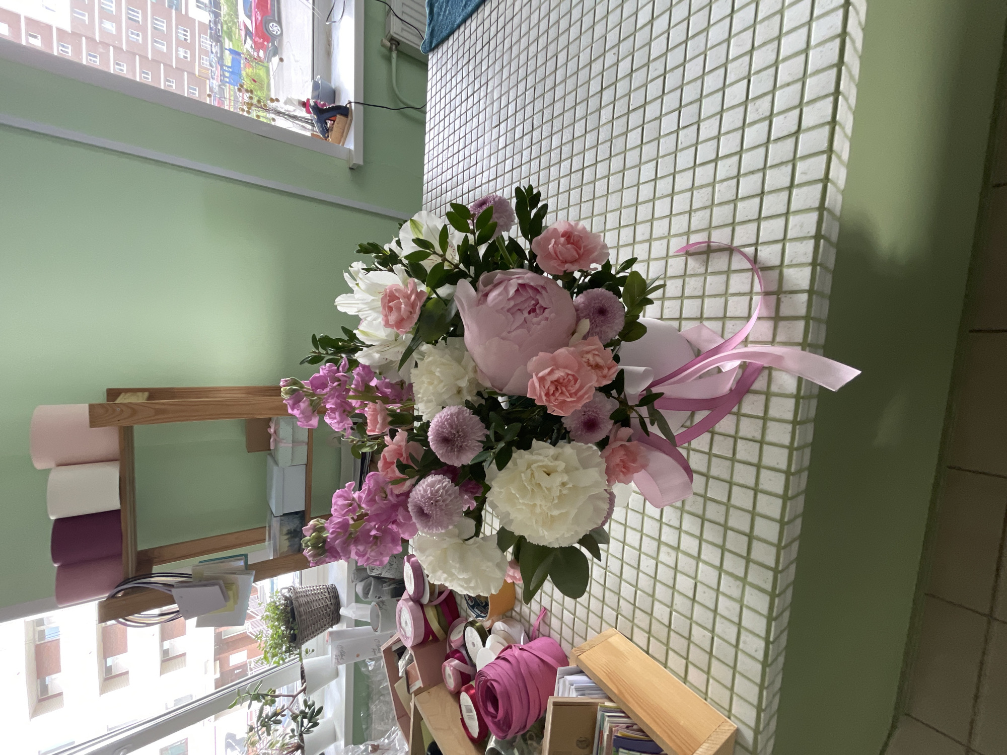 A high-angle, indoor shot shows a bouquet of flowers with a pink ribbon in an unknown location. The bouquet is in the center of the frame, comprised of pink, purple, and white flowers with green leaves. A large pink peony is centered in the bouquet. A light pink ribbon trails from the right side of the bouquet.
Behind the bouquet, the wall is covered in small, white square tiles. To the left of the bouquet, the wall is light green. Below the bouquet, a shelf holds rolls of pink, white, and purple paper. Other items on the shelf include a basket, ribbon rolls, and paper stacks.
A window is in the background on the left side, showing a building with several windows. The image is rotated counterclockwise, making the scene appear to be on its side.