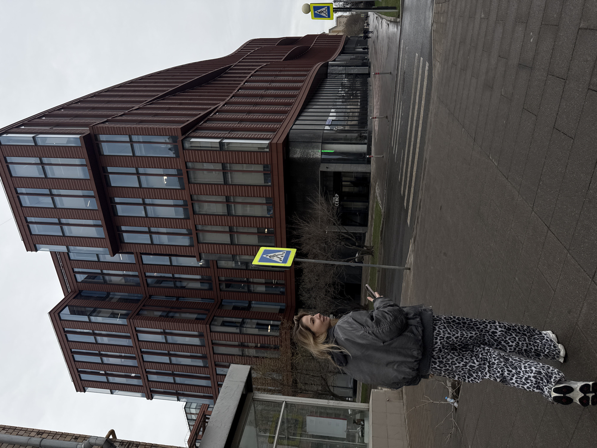 A woman with blonde hair wearing a black jacket and patterned pants stands on a sidewalk next to a building. She is looking at a phone she is holding. Behind her is a modern architectural building with many windows, appearing to be a commercial or office structure. In the background, a pedestrian crosswalk sign is visible. The scene is outdoors and the sky is overcast. Based on the architecture and signage, the location is in Moscow, Russia.
