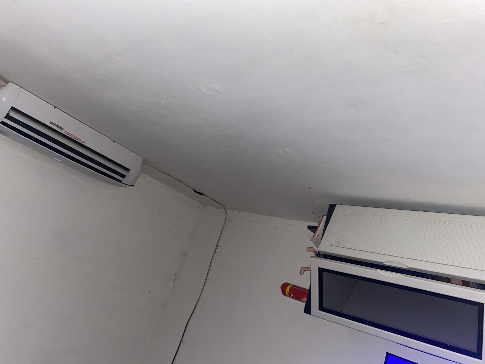 This image depicts an indoor scene, likely a room in a residential or commercial building in South Tangerang, Indonesia. Dominating the upper portion of the frame is a white air conditioning unit mounted on a white wall. Below and to the left, two white cabinets are visible. The cabinet on the left has a dark, reflective surface, possibly a television screen or a glass-fronted cabinet. The cabinet on the right features a textured white door. On top of these cabinets, several small objects are present, including a red cylindrical item, a red and white bottle, and some small figurine-like objects with pale skin tones. Wires run along the ceiling in the upper left quadrant of the image. The walls appear to be painted white and show some signs of wear, with minor cracks and discoloration visible. The lighting suggests an indoor environment, possibly with artificial light sources. The perspective is slightly tilted, looking upwards towards the air conditioning unit and the ceiling.