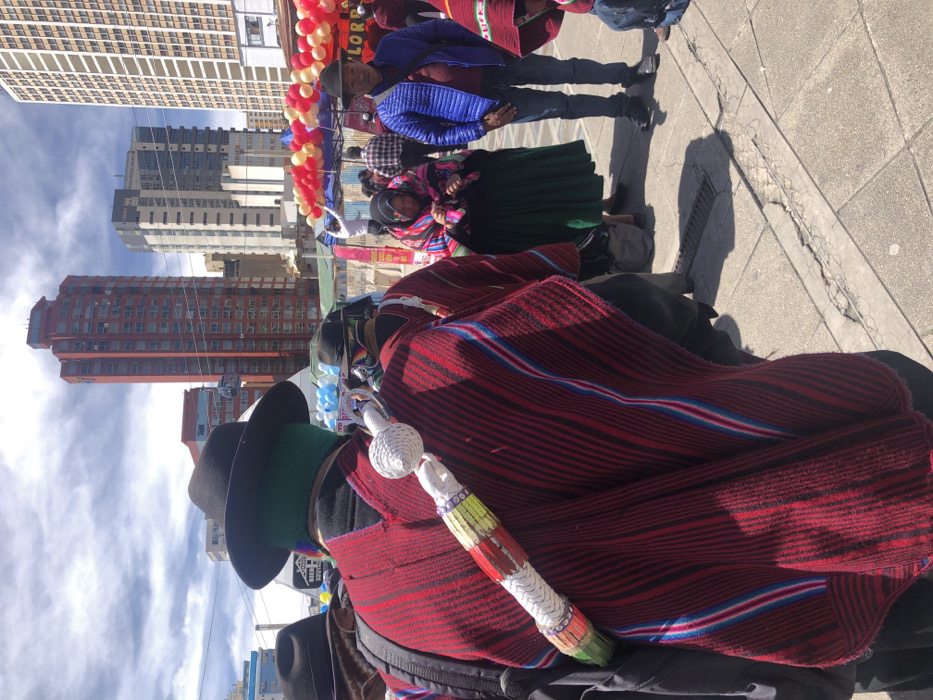 The image depicts an outdoor urban scene on a bright, sunny day, identified as El Prado avenue in La Paz, Bolivia. The foreground is dominated by the back of an individual wearing a dark-brimmed hat, a green head covering, and a striped red poncho with black, blue, and light red accents. A white, woven, club-like object, adorned with yellow, red, and green patterns, is strapped diagonally across this person's back. To their right, another individual in a dark hat and red poncho is partially visible.
In the mid-ground, a woman with dark hair wears a colorful striped manta over a dark top and a dark green pleated skirt, looking downwards. Adjacent to her stands a man in a blue puffer jacket, jeans, and a dark hat, facing towards the right. Overhead, red, yellow, and white balloons are strung across the scene, indicating a festive event or market stalls. Further left, blue and white balloons are visible. Temporary structures or tents are present behind these individuals.
The background features the distinct, tall, red-colored Torre Girasoles building, prominent among several other modern high-rise buildings of varying grey and white tones that characterize the urban skyline. A cable and support structure from La Paz's Mi Teleférico system is visible in the upper left. The ground is a paved pedestrian area with visible cracks and a drainage grate, casting sharp shadows from the people, indicating strong sunlight. Multiple other indistinct figures are present in the deeper background, with one individual on a structure appearing to work with the balloons.