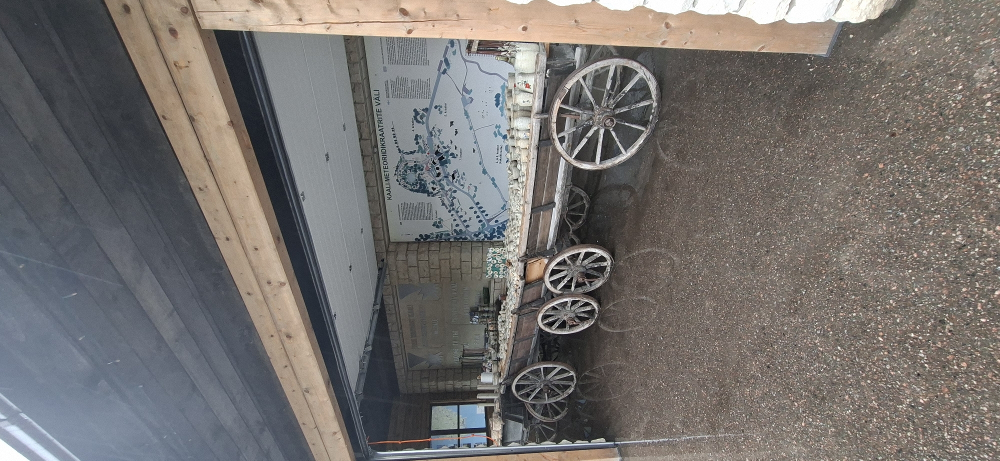 The image shows a long, antique wooden wagon filled with numerous small white pots and jars. The wagon has several large spoked wheels. It is positioned in front of a brick wall displaying a large map or diagram with text in Estonian, titled "KAALI METEORIIDIKRAATRITE VÄLI." The wagon and its contents are placed on a wet, textured stone floor. The scene appears to be in an open-air pavilion or covered outdoor area, with wooden beams visible overhead and to the right. The setting is the Kaali meteorite crater field in Estonia, likely a visitor center or exhibition area. There are no people visible in the image.