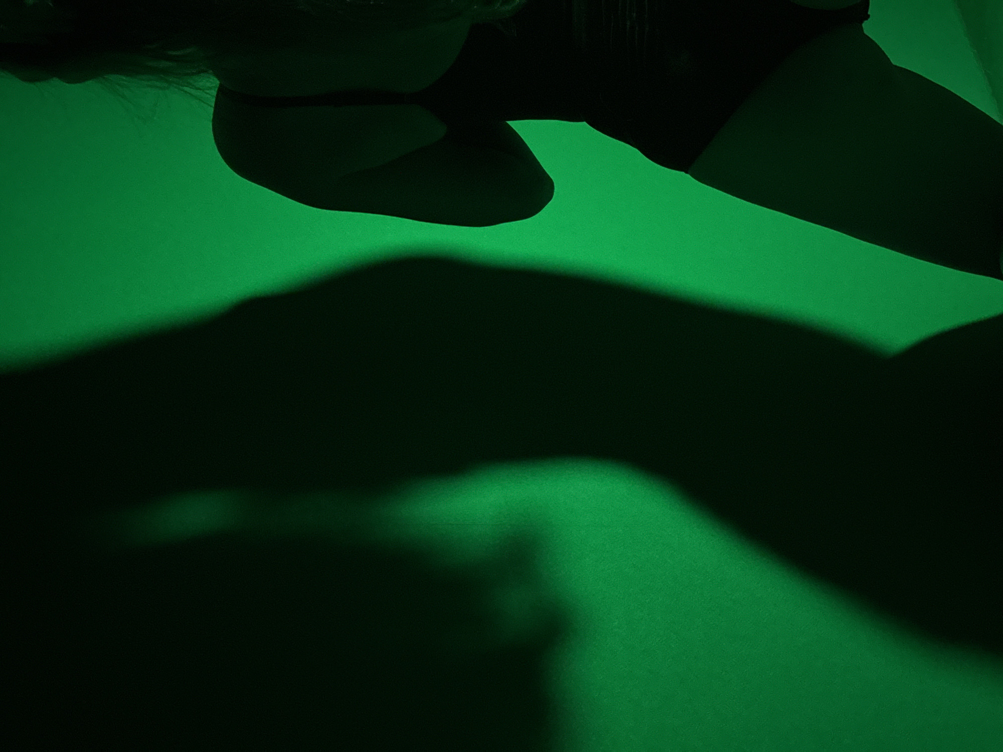 The image shows a person, apparently female, lying down in dim, greenish lighting. Only the upper body and part of the legs are visible. The person wears dark clothing. A shadow is cast on what appears to be a surface, possibly a floor or wall, also illuminated in green. There is no indication of the location being Chaikovsky, Russia, or any city for that matter.