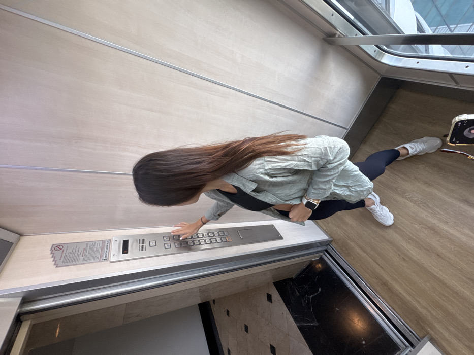 The image captures a high-angle view of a female individual in an elevator lobby within a modern building in Bangkok, Thailand. The subject, with long, dark brown hair, is positioned facing an elevator control panel on the left. She wears a black crop top, an unbuttoned light green/grey long-sleeved shirt, dark leggings, and white athletic shoes. A white smartwatch and gold-colored bracelets are visible on her left wrist. Her right hand's index finger is extended, pressing a button on the stainless steel elevator panel, which displays the letter 'P'. Above the control panel is a rectangular sign featuring Thai text and the company name "STANDARD ELEVATOR SERVICE CO.,LTD." The elevator door to the left is open, revealing a dark, reflective floor inside and a black and white tiled pattern on the floor beyond the threshold. The immediate flooring is a light wood-look material, contrasting with the light wood-paneled walls accented by vertical metallic strips. A smartphone with its screen visible is placed on the floor in the bottom right corner of the frame. Through a large window on the right, modern high-rise building exteriors with reflective glass facades are partially visible, indicative of an urban environment.