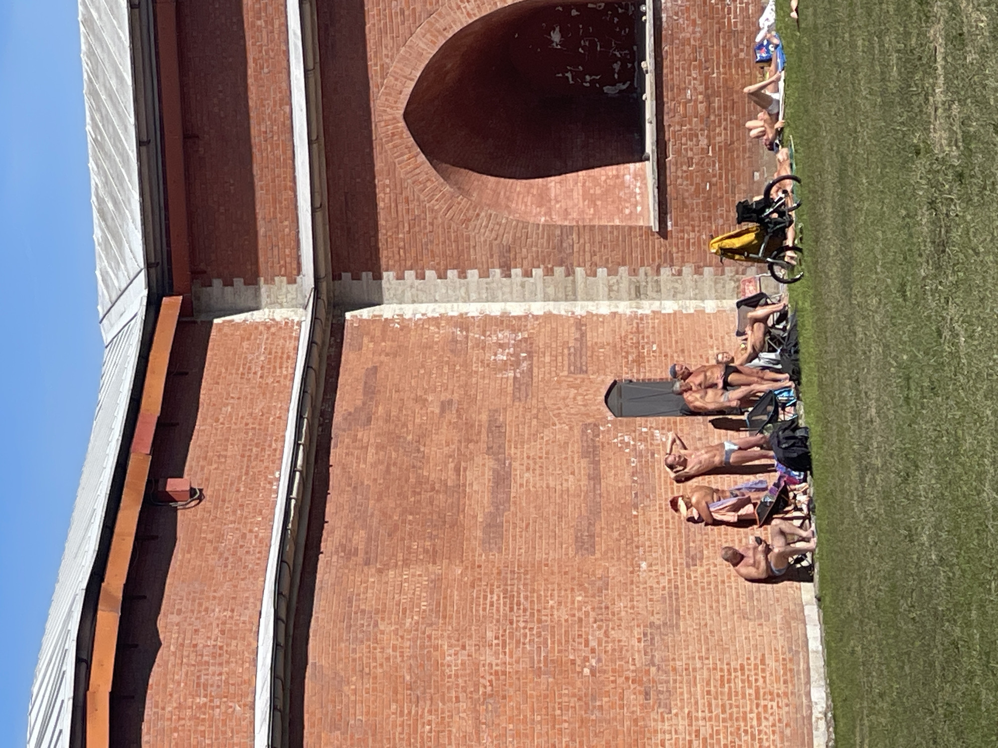 The image depicts a brick building with a large arched opening near its top. A grassy area stretches out beside the building. Several individuals are sunbathing next to the building. A bicycle with a yellow bag attached leans against the building further down. The location is Saint Petersburg, Russia, as evidenced by the architecture of the building.