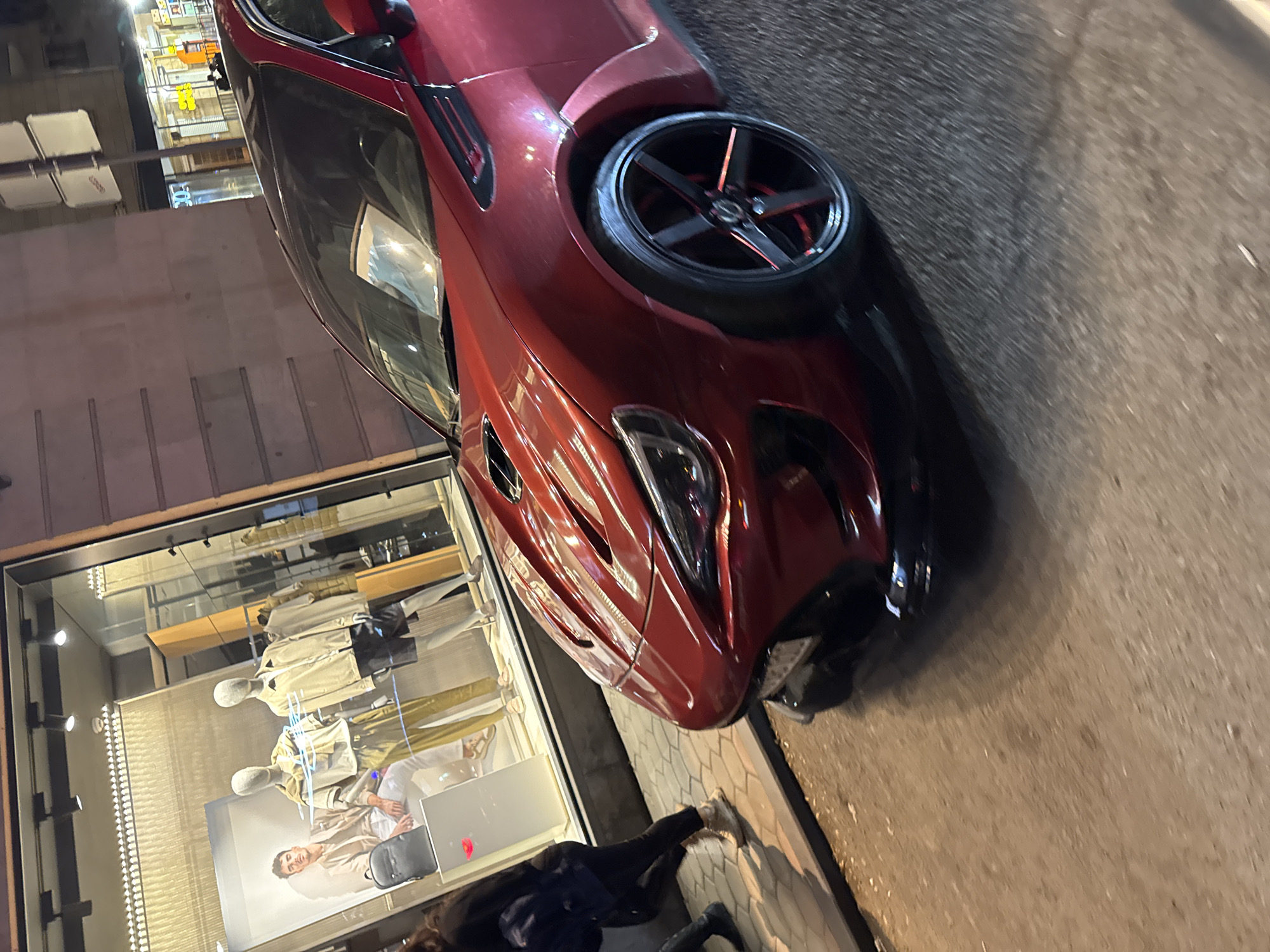 The image, rotated 90 degrees clockwise, captures a vibrant night scene in an urban area of Yerevan, Armenia. A sleek, dark red sports car, possibly a coupe, is prominently parked on a patterned sidewalk. The car features custom black multi-spoke wheels with subtle red accents, and its highly reflective surface mirrors the surrounding lights. The front end of the vehicle, including a headlight and grille, is clearly visible.
Adjacent to the car is a well-lit glass storefront, showcasing several mannequins dressed in contemporary fashion, primarily in beige and light earth tones. A large visual display inside the store features a smiling man. At the bottom edge of the frame, the lower body of a person, dressed in dark clothing and white sneakers, is visible, standing near the storefront. The pavement beneath the car and the pedestrian area is distinctly patterned, while the surface beyond the car appears to be a darker, rougher street. The overall impression is that of a bustling city street or shopping district after dark, with artificial lights illuminating the scene. No discernible text is clearly legible in the image, though a faint logo on the car's wheel hub and some unreadable text on the store's display can be observed.