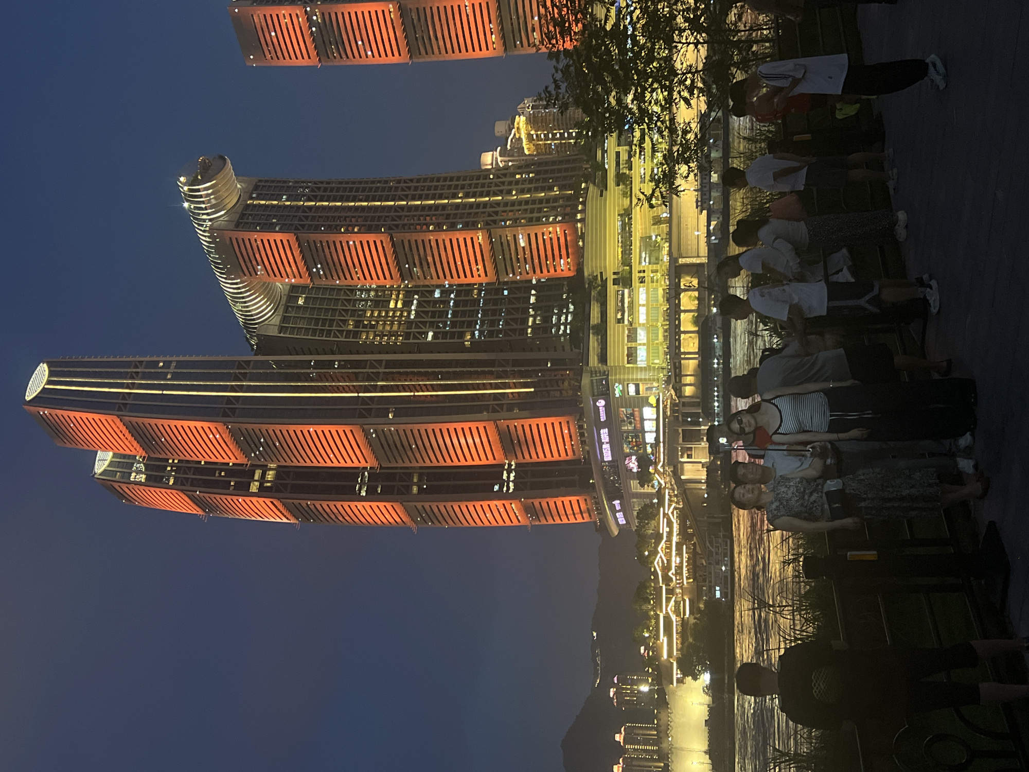 In the Yuzhong District of China, at dusk, a group of people stands by a riverfront promenade, facing illuminated skyscrapers. The buildings, characterized by their orange and yellow lighting, dominate the background. One skyscraper features a distinct, curved, horizontal element near its top, resembling a bridge or a ring. Another skyscraper is composed of twin towers. The foreground shows a railing and a dark, paved area where the individuals are gathered. The people are casually dressed, with some taking photos or looking at their phones. A river flows between the promenade and the buildings, with boats visible on its surface. The sky is a deep blue, indicative of twilight.