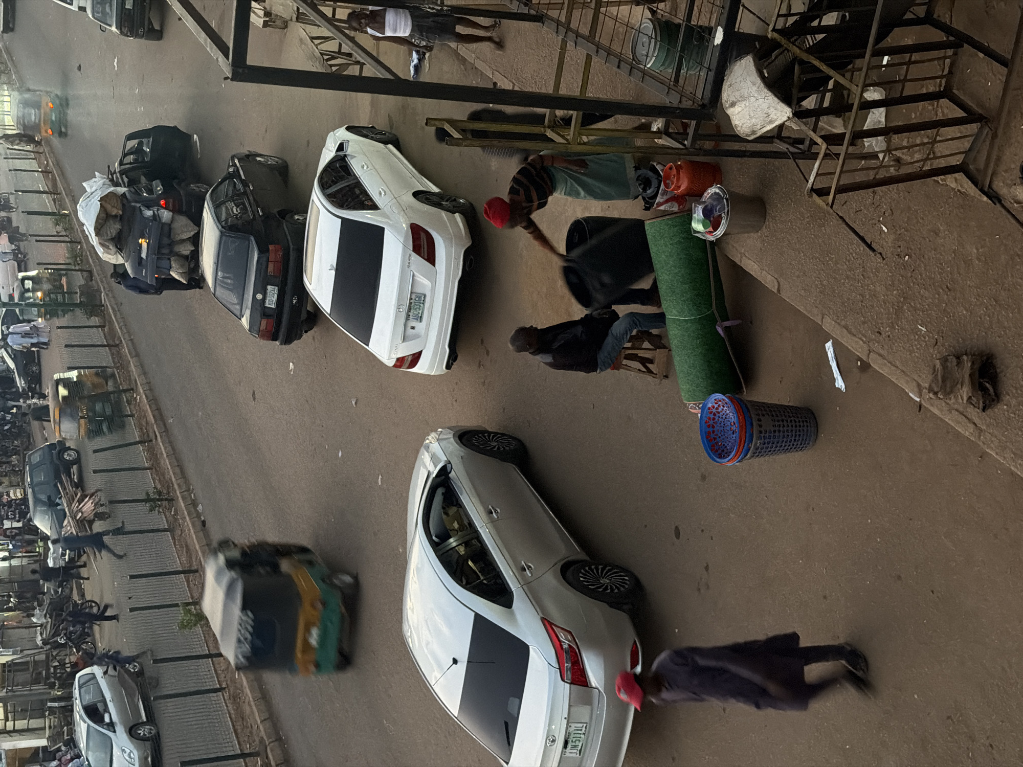 The image depicts a street scene in Bukuru, Nigeria, with a focus on transportation and commerce. Several vehicles are visible, including a silver sedan, a white sedan, a black SUV with a heavily loaded roof rack, and a three-wheeled auto-rickshaw. Pedestrians are present, some walking along the roadside and others interacting with objects on the ground. Two individuals are near a collection of large black containers, one of whom appears to be lifting or moving a container. Another person, wearing a red cap, walks in the foreground, blurred due to motion. The environment suggests a bustling urban setting with a paved road, sidewalks, and a market or roadside stall area with various items like stacked containers, a rolled-up green material, and a gas cylinder. The time of day appears to be daytime, given the lighting conditions.