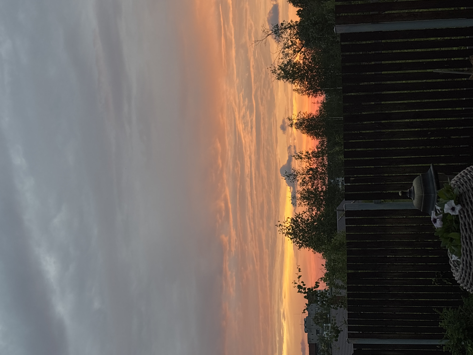 The image depicts a sunset over a residential area. In the foreground, a dark wooden fence stretches horizontally across the frame, with a woven basket containing white flowers and a lamppost to its right. Beyond the fence, a line of trees with green foliage forms a natural barrier. In the background, buildings and rooftops are visible, suggesting an urban or suburban setting. The sky is filled with clouds, with hues of orange, yellow, and gray indicating the time of day is sunset. The overall scene is peaceful and serene. The location is the city of Gubakha, Russia.