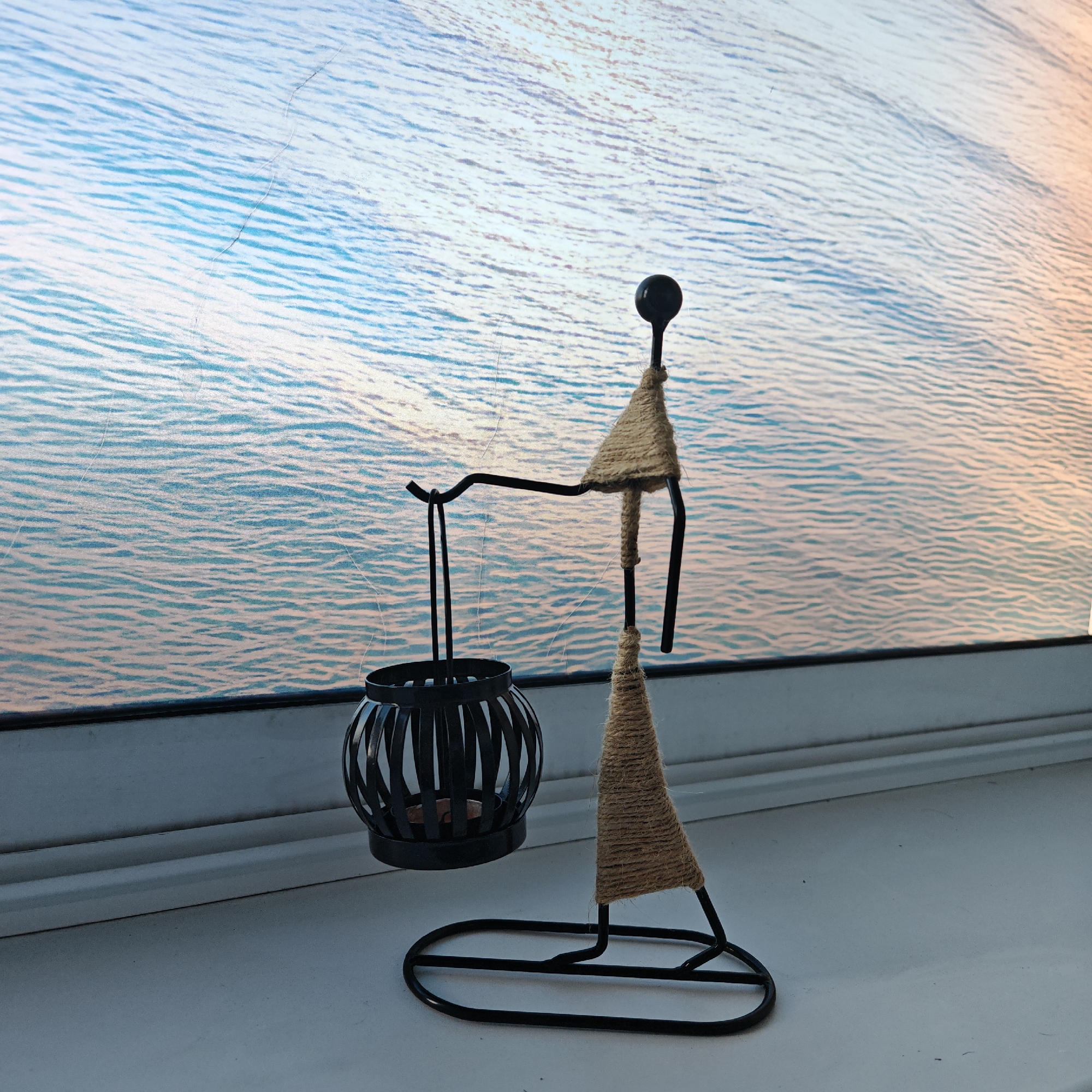 The image shows a decorative metal candle holder, shaped like a figure carrying a lantern, on a windowsill. The candle holder is black metal with sections wrapped in natural fiber twine. Behind the candle holder is a window or surface with a pattern resembling water. There is no evidence suggesting the image was taken in Rostov, Russia. The scene is indoors and appears to be a still life setup. No people are visible.