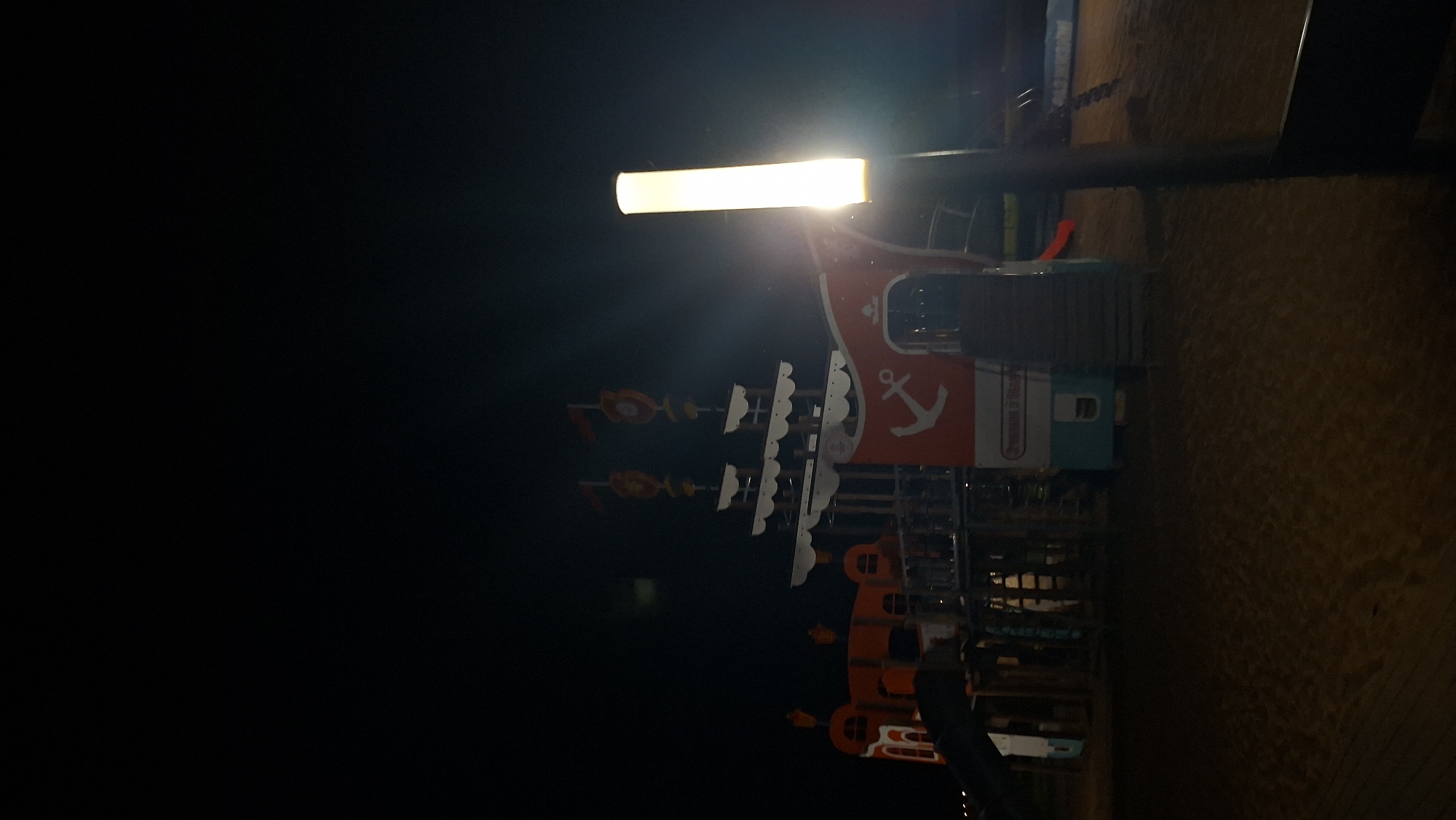 This is a nighttime photograph of a playground designed to resemble a pirate ship. The structure is brightly colored with red and white accents and features two masts with flags. An anchor symbol is prominently displayed on the side of the ship. To the right of the playground, a tall, illuminated light post stands, casting a bright glow on the scene. The playground is situated on a sandy surface, suggesting it might be located near a beach or a sand-covered recreational area. The overall impression is one of a well-lit, albeit empty, play area after dark. There is no indication of the specific location within the city of Myadzyel, Belarus.