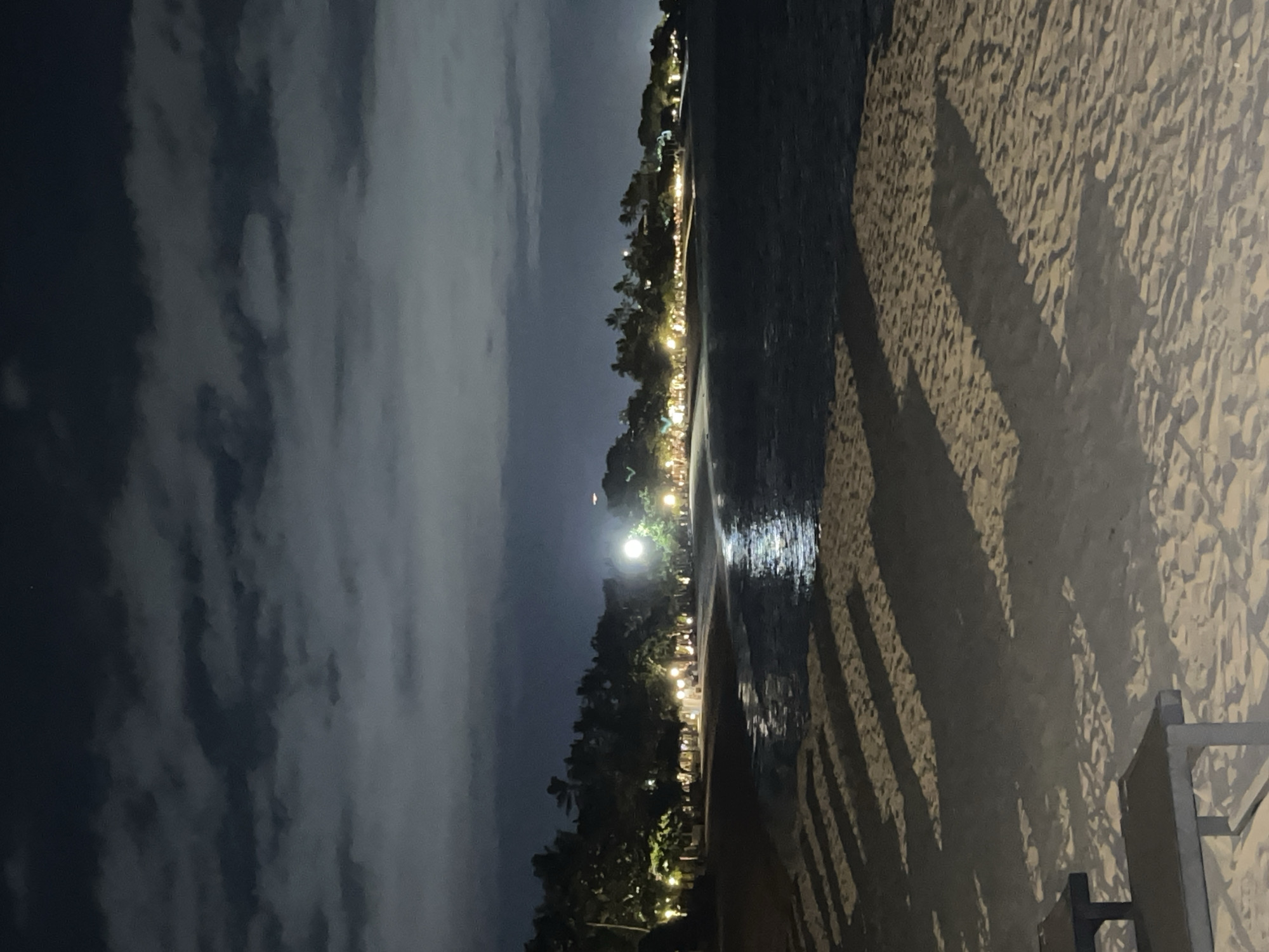 A night-time view of a beach near Unknown, Indonesia. The sky is cloudy, with darkness prevailing on the left and lighter areas with cloud detail on the right. Along the horizon, lights from buildings and structures are visible, indicating a populated area. A line of trees separates the lights from the beach. The beach slopes downwards to the sea. The sea reflects light from the shore, creating a shimmering effect. The sand of the beach is light in color, and shadows, possibly cast by a structure, are visible across it. A partial view of what appears to be a bench or similar structure is visible in the lower right corner. No people are visible in the image. The environment is coastal and dimly lit.