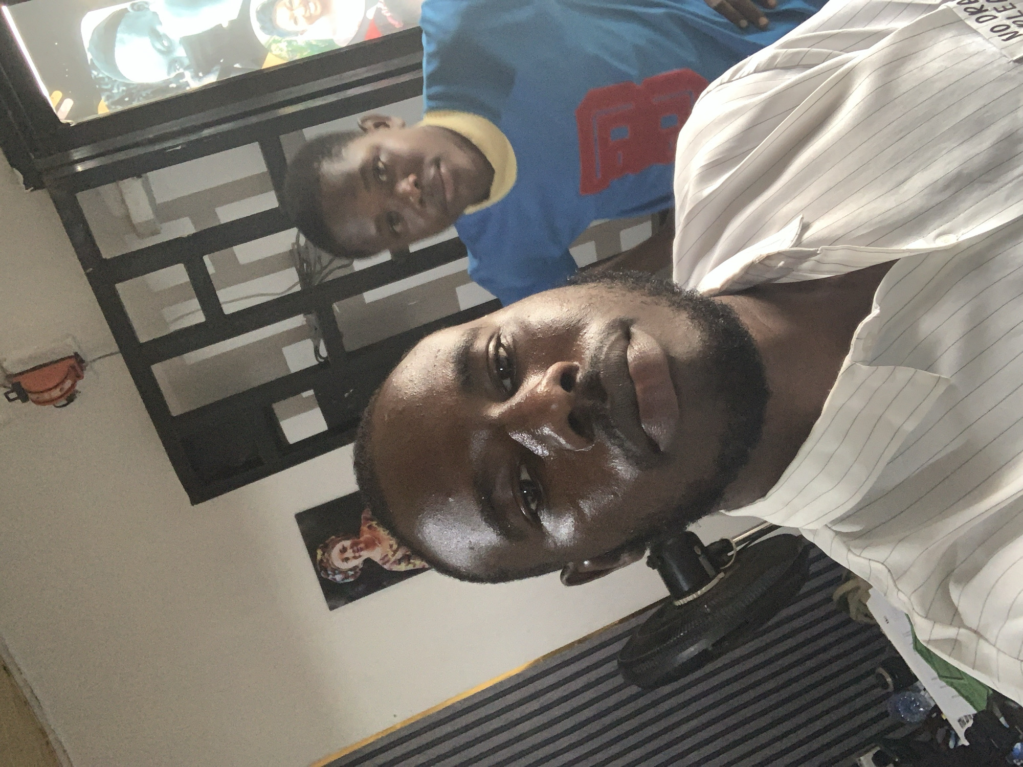 The image shows two people in a room. One person is wearing a white striped shirt. The other person is wearing a blue shirt with a red number. There is a television in the background with people on the screen. There is also a black fan and other objects in the room. No location in Jos, Nigeria can be determined from this image.