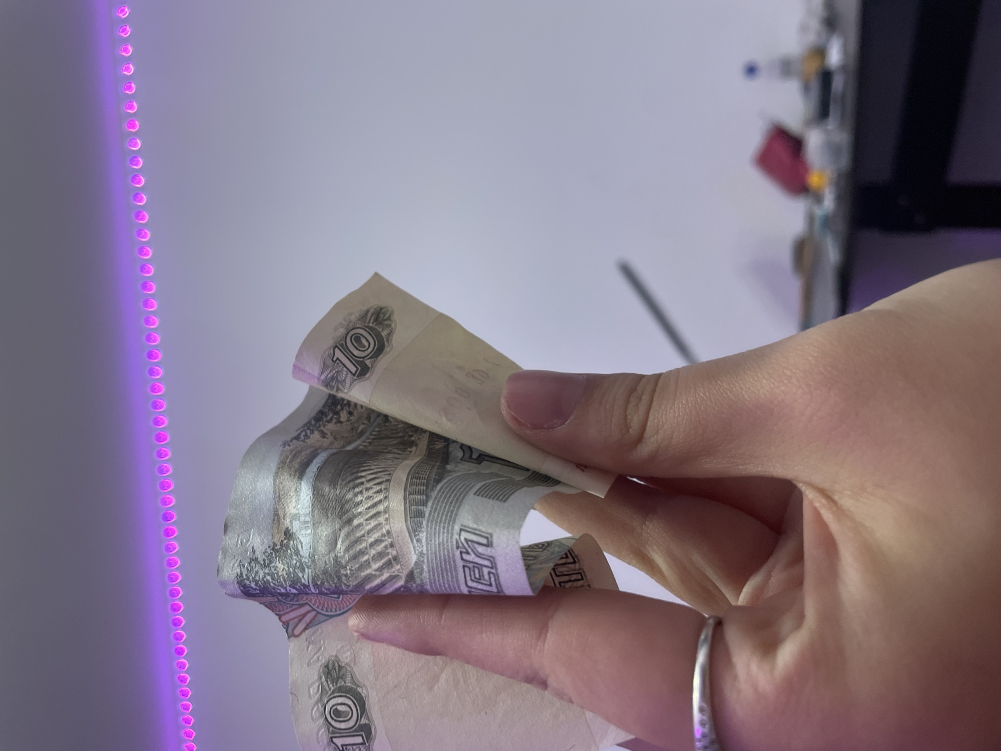 A person's hand, wearing a silver ring on their left ring finger, holds crumpled Russian 10 ruble banknotes. The banknotes are predominantly light brown and cream with dark grey and black printing depicting architectural elements and the numeral "10." The hand is positioned in the foreground, obscuring a portion of the banknotes. The background features a white wall illuminated by a string of pink LED lights that form a horizontal line across the top of the image. Below the wall, a dark surface, possibly a desk or table, is visible, cluttered with out-of-focus objects including a pink item, a bottle, and other indistinct items. The scene is likely indoors, potentially in an apartment or office setting, in Kokkorevo, Russia.