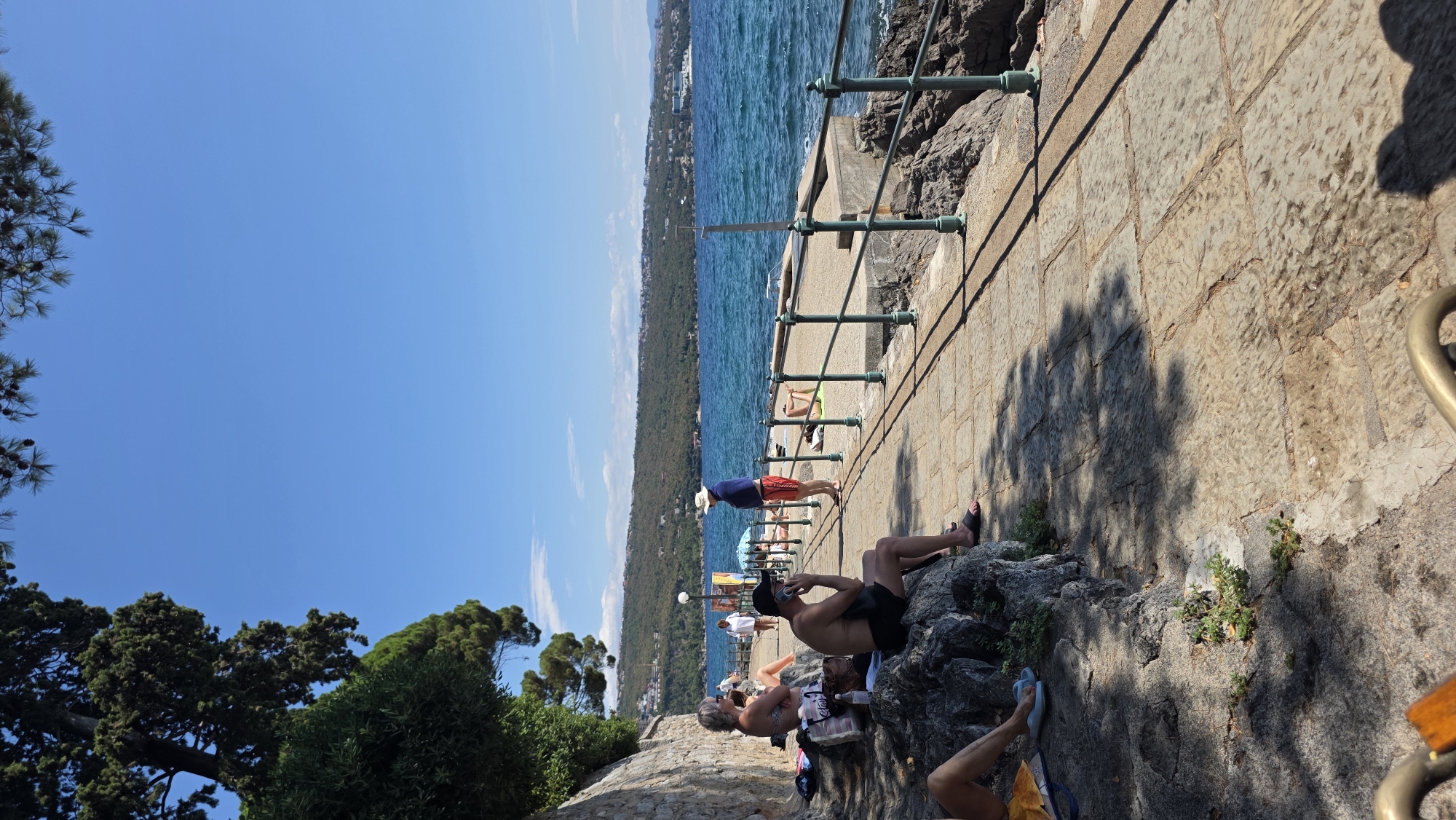 The image depicts a sunny day along the coast of Grad Opatija, Croatia, likely on the Lungomare, a famous promenade. In the foreground, several people are relaxing on a stone embankment by the sea. One man in black swim trunks is sitting on rocks, talking on his phone. To his left, a woman in a bikini sits on the rocks with her legs outstretched. Further down the promenade, a man in red shorts and a blue towel draped over his shoulders stands and looks out at the sea. Several other individuals are visible sunbathing on towels or sitting on the promenade. The turquoise sea is to the right, with waves gently lapping against the rocky shore. A stone walkway with a metal railing lines the promenade. In the background, a lush green hillside dotted with buildings stretches along the coast, and the blue sky is clear with a few scattered clouds. A large ship is visible in the distance on the water. Trees and vegetation are visible on the left side of the image, casting shadows on the ground.