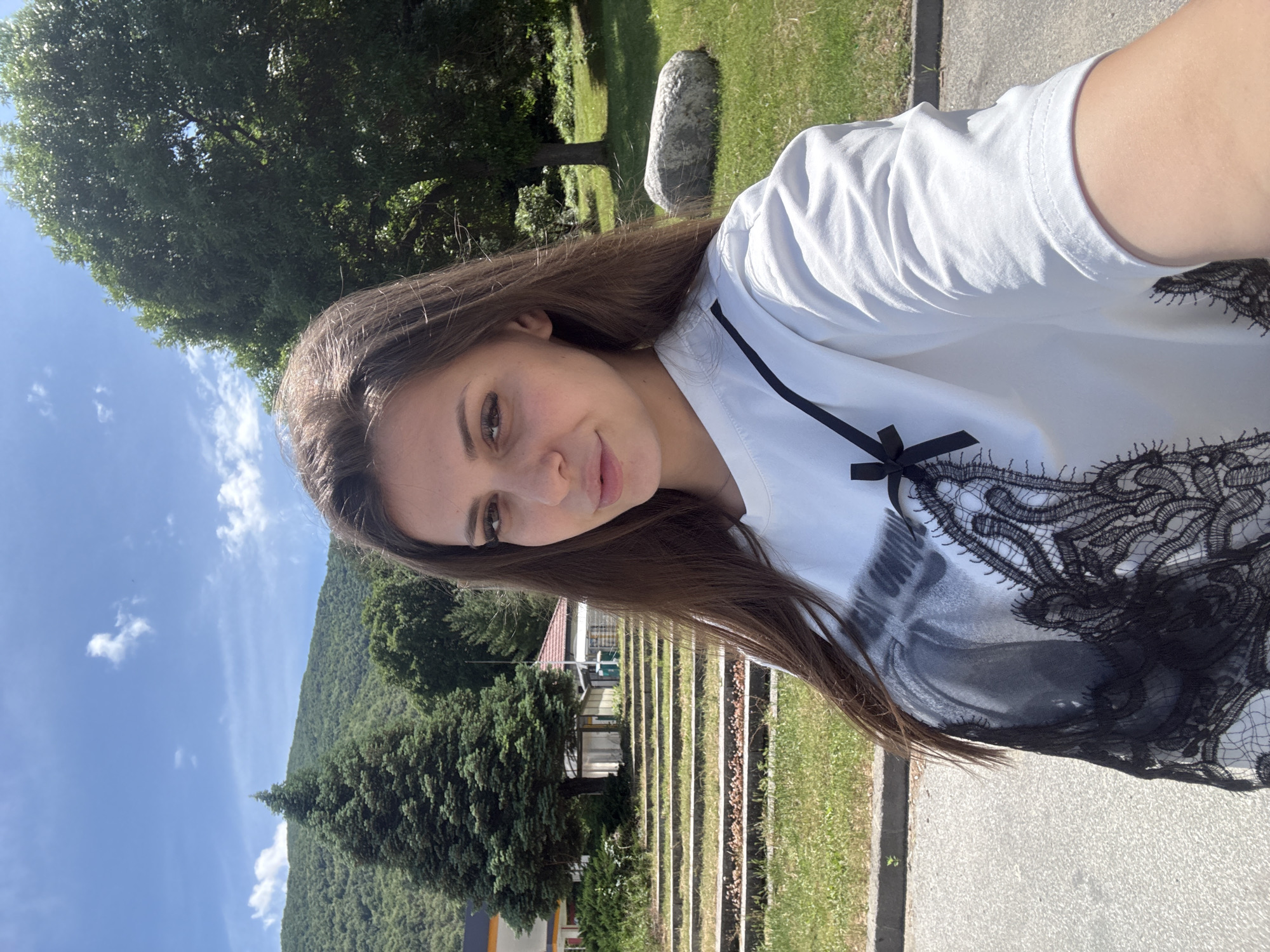 The image is a bright, sunlit selfie taken outdoors, featuring a young woman against a natural and rural backdrop. The photograph is rotated 90 degrees clockwise, presenting the scene sideways.
The primary subject is a young woman, shown from the chest up. She has long, straight, dark brown hair, a fair complexion, and a gentle smile. Her dark eyes are directed towards the camera. She is wearing a white t-shirt that features a prominent black lace pattern across the chest and shoulder area, accompanied by a small black ribbon or bow near the left shoulder.
The setting is a picturesque outdoor environment. The sky is a clear, vibrant blue with a few scattered white clouds, suggesting a sunny daytime. In the background, a large, green, tree-covered mountain or hill dominates the horizon. Various types of trees, including large deciduous trees with full green canopies and darker coniferous trees, are visible throughout the mid-ground and background.
To the woman's right, there is a paved path or road next to a grassy area, where a large, light grey rock is situated. To her left, behind some foliage, part of a building with a red roof can be seen. Below this building, there are distinct terraced structures or steps, possibly for agricultural or landscaping purposes, extending across the mid-ground. The overall impression is one of peace and natural beauty. There is no discernible text visible in the image. The location context indicates this scene is in Vetren Dol, Bulgaria.