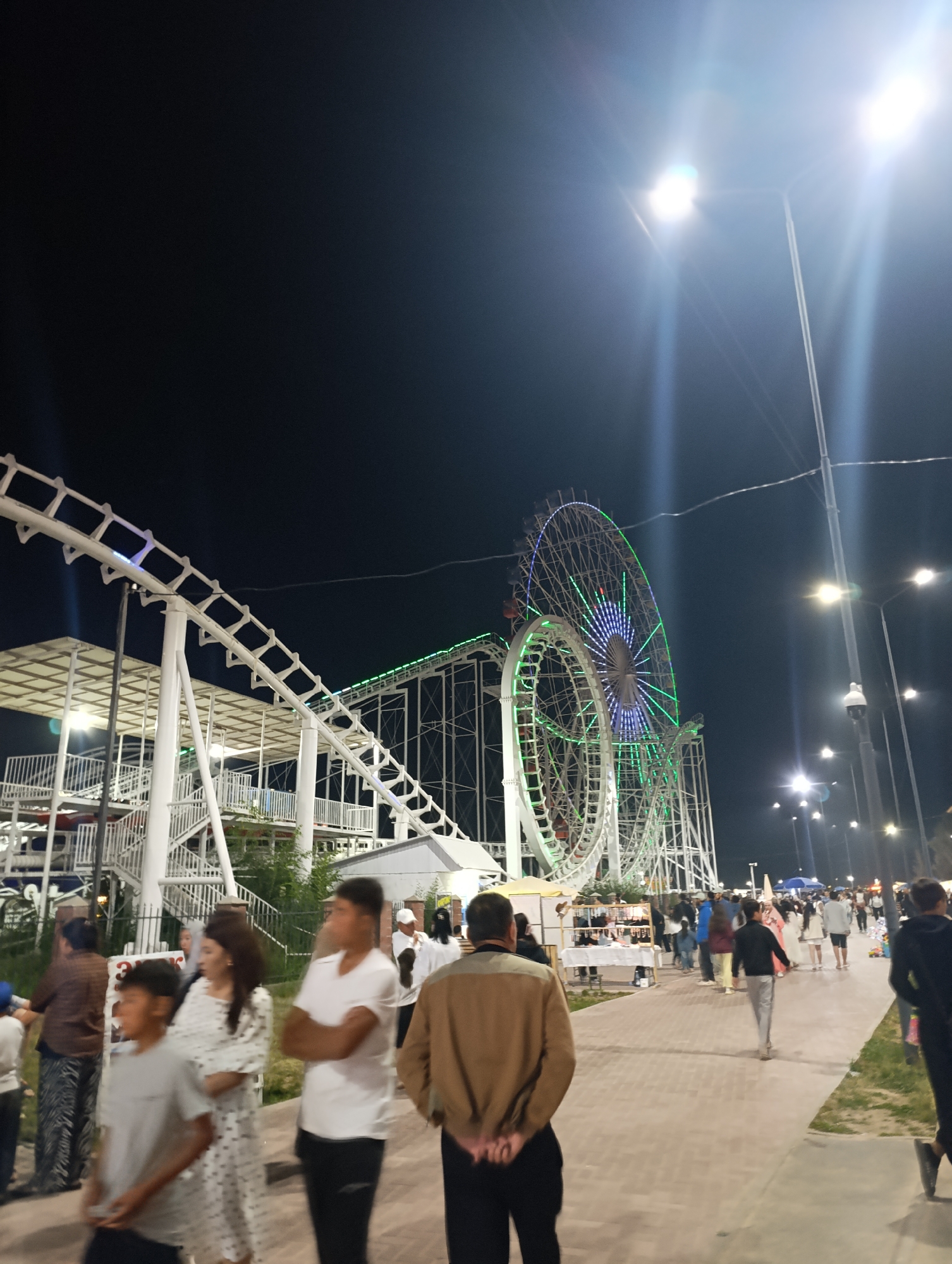 This nighttime, outdoor photograph depicts an amusement park scene in Bosteri, Kyrgyzstan, near the shores of Issyk-Kul Lake. A large Ferris wheel, illuminated with green and blue lights, dominates the background, along with a roller coaster structure. Several street lamps, also brightly lit, illuminate the area.
In the foreground and midground, a crowd of people is visible. Many are walking along a paved pathway, while others are gathered near stalls or attractions. The individuals appear to be of various ages and are dressed in casual attire. Some are engaged in conversation, while others are simply strolling. The overall atmosphere suggests a lively and active evening at the amusement park. The ground is paved with cobblestones, and there is a strip of grass to the right of the path. Some of the people are out of focus due to the motion blur.