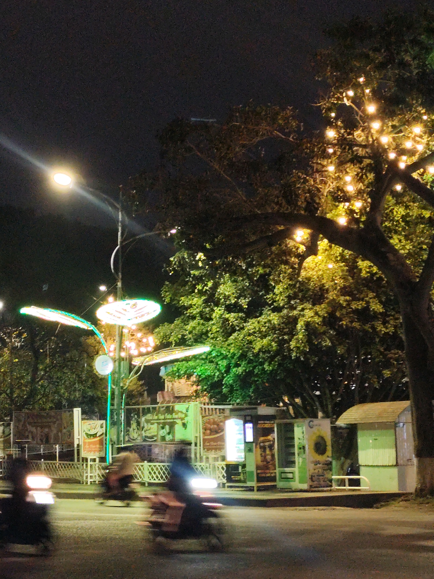 This night scene depicts a lively street in Da Lat, Vietnam, under a dark, clear sky. Artificial lights are the primary source of illumination. A bright streetlight in the upper left casts a strong beam, while a large tree on the right is adorned with numerous small, warm-toned string lights, creating a festive glow.
In the center, a tall pole supports unique, glowing decorative light structures shaped like circles and wings, emitting green, white, and pink light. The paved road below shows several blurred figures on motorcycles, their streaked headlights and taillights indicating active evening traffic and motion.
Along the right side of the street, there are multiple vending machines and small kiosks. One vending machine prominently features a large sunflower image. Another machine next to it has illuminated product advertisements, with some indistinct text, possibly including the word "CHICKEN." Further to the right, a small, light-colored wooden stall or kiosk is visible, along with white metal barriers or fences behind these roadside establishments. The scene conveys a bustling urban evening atmosphere.