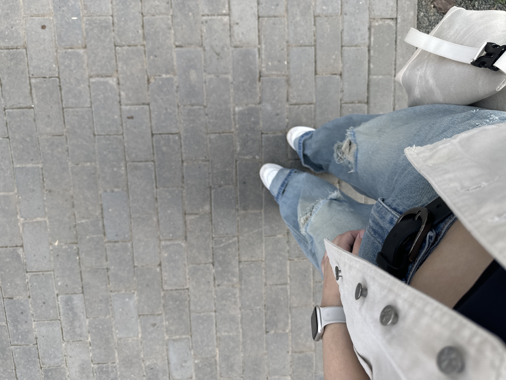 A first-person shot shows a person standing on a grey brick sidewalk. The individual is wearing ripped light blue jeans, white sneakers with light grey socks, and a white denim jacket. A black belt is visible. A white smartwatch is on the person's left wrist. Part of a white tote bag with a white strap is visible in the upper right corner of the image. The location is identified as Perm, Russia, based on image analysis. The photograph focuses on the person's attire and the surrounding urban pavement.