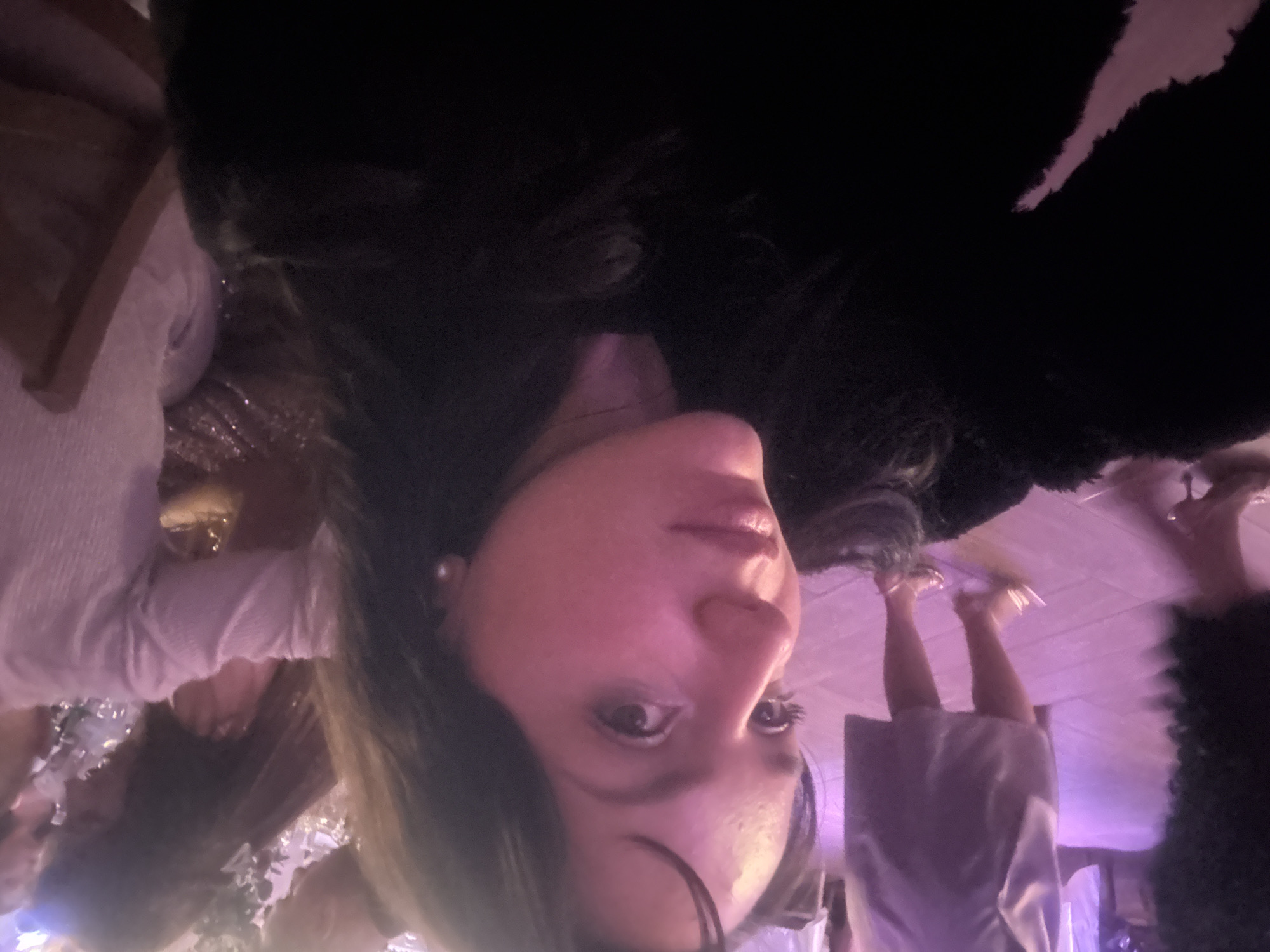 A close-up, rotated view depicts a woman with dark hair, fair skin, and visible eyes. She wears a light-colored top. A dark, possibly furry object is above her head. A group of people are present in the periphery of the image, some wearing lighter colors. The background suggests an indoor environment. The location is not discernible from the visual information provided.