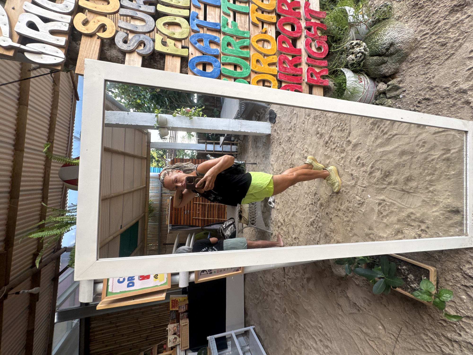 The image shows a woman taking a selfie in a mirror outside a building in Kuta, Indonesia. The mirror is leaning against a wall. The woman is wearing a black top and neon green shorts, along with yellow sneakers. She is holding a phone and taking a picture of herself. In the reflection, another person is visible lying on a lounger, appearing to relax. The background includes a sign with colorful letters, plants in pots, and the sandy ground characteristic of the area. The building is a shop.