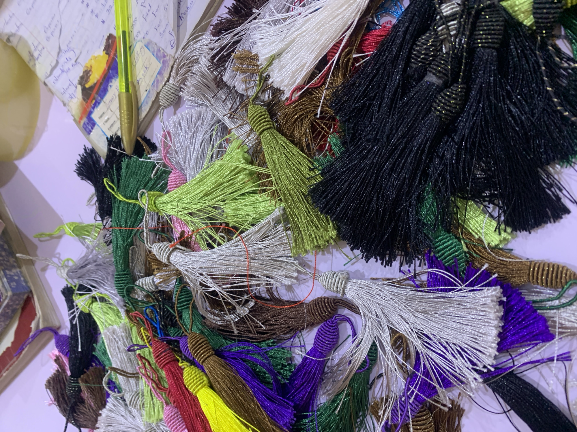 This image depicts a close-up view of a collection of colorful tassels scattered on a light purple surface. The tassels are made of thin, fibrous threads and vary in color, including black, white, silver, gold, green, purple, red, and brown. Some tassels are neatly bundled, while others are loose and intertwined. In the background, a stack of books and a yellow pen are visible, suggesting a craft or study area. The lighting is somewhat dim, creating a warm and slightly diffused atmosphere. There are no people visible in the image. The specific location in Gura Top, Nigeria, cannot be determined from the visual information provided in the image.