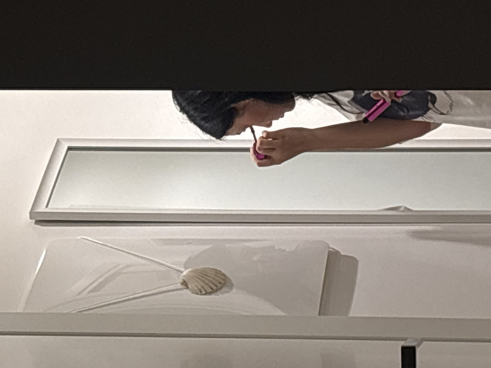 The image captures a young woman applying makeup in a bathroom. She is standing in front of a mirror, holding a pink makeup applicator to her eye. Her dark hair is visible as is part of her t-shirt, which has a graphic design on it. To the left of the mirror, a framed picture of a seashell hangs on a white wall. The scene appears to be indoors, possibly in a residential setting in Cupra Marittima, Italy, as suggested by the white walls and the framed artwork. The overall lighting is soft, creating gentle shadows and highlighting the woman's focused expression.