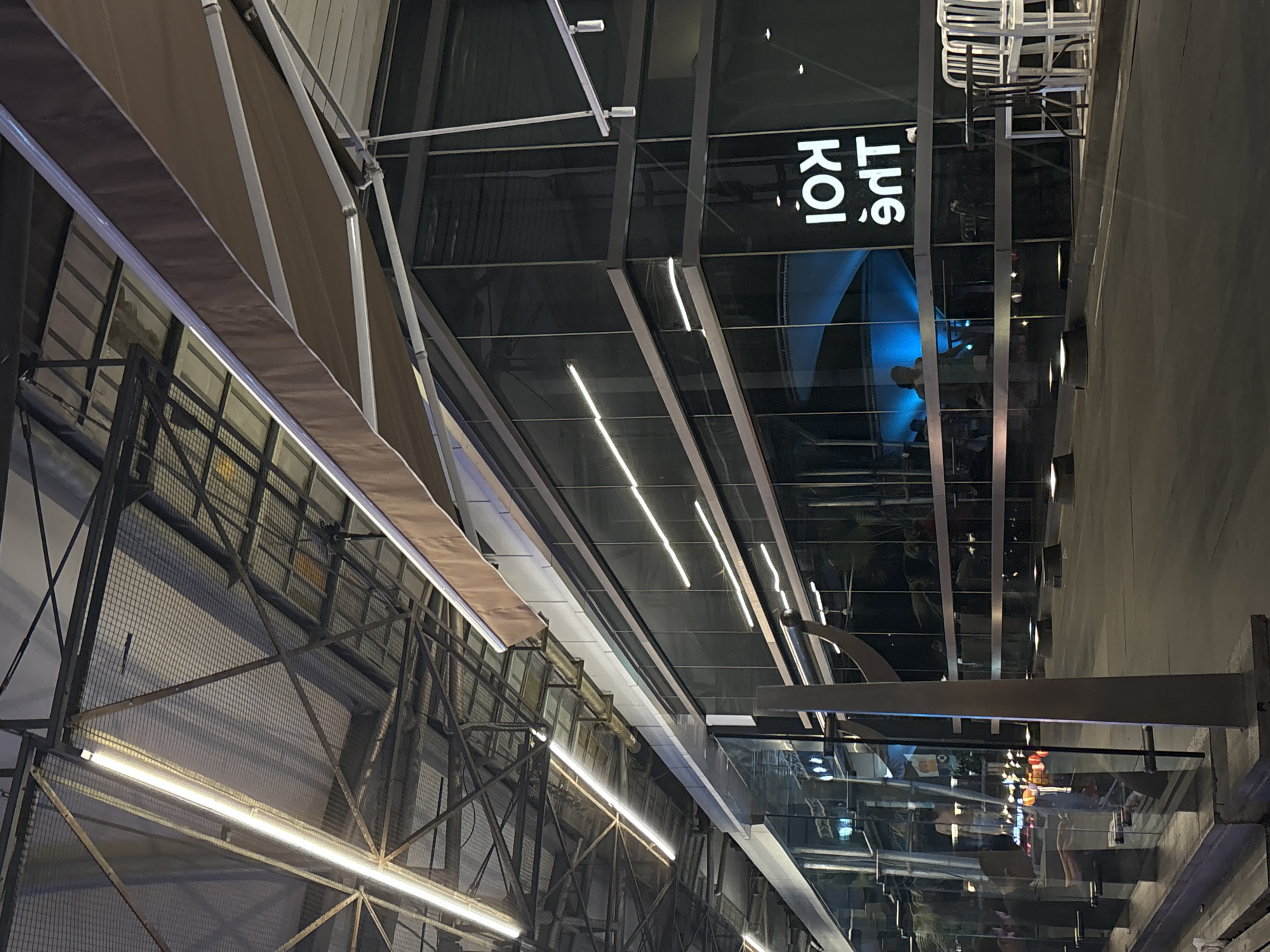 This is an outdoor, dimly lit scene in North Jakarta, Indonesia, featuring a restaurant or business named "Koi". The building has a modern architectural design, characterized by extensive use of glass and metal framework. A series of awnings extend from the side of the building. The metal framework includes a wire mesh structure above the awnings, with integrated lighting fixtures. A blue light illuminates a section of the building's interior. The scene also features stacked white chairs.