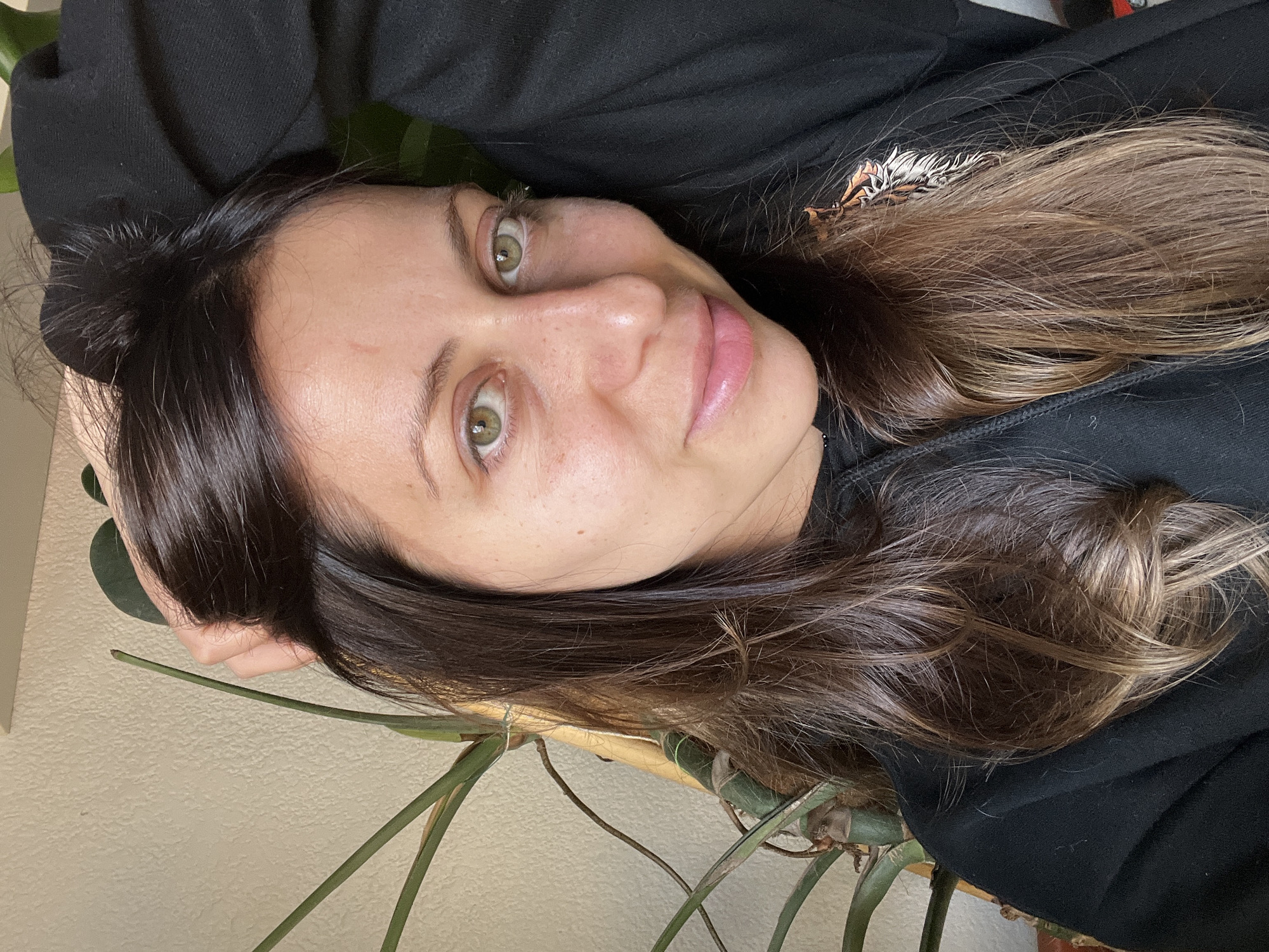 A close-up portrait features a woman with fair skin, striking green eyes, and long, dark brown hair subtly highlighted with lighter brown tones. She is reclining and looking directly at the viewer with a calm, engaging expression, her lips slightly parted with a light pink hue. She wears a black hooded sweatshirt, with a small decorative pattern, possibly animal print, visible on the right side. The background includes large green plant leaves and stems, suggesting an indoor setting with a houseplant, and a light-colored, textured wall. The scene is illuminated by soft, natural daylight, creating gentle highlights on her hair and skin, indicating a daytime shot. While no specific location cues are visible in the image, the context provided states the location is Barcelona, Spain. No visible text is present.