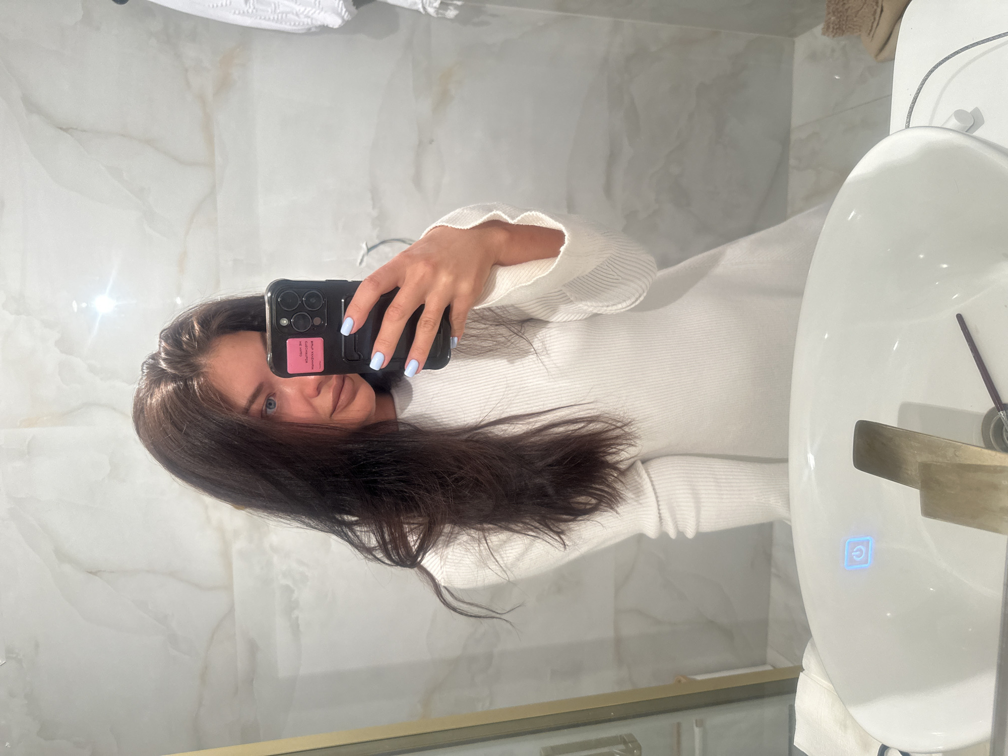 A woman with dark hair is taking a selfie in a bathroom. She is holding a black smartphone with a pink sticker on the back and has light blue nail polish. She is wearing a white sweater. The bathroom has marble walls and a white sink with a gold-colored faucet. The location is not specified in the image and cannot be determined to be Moscow, Russia.
