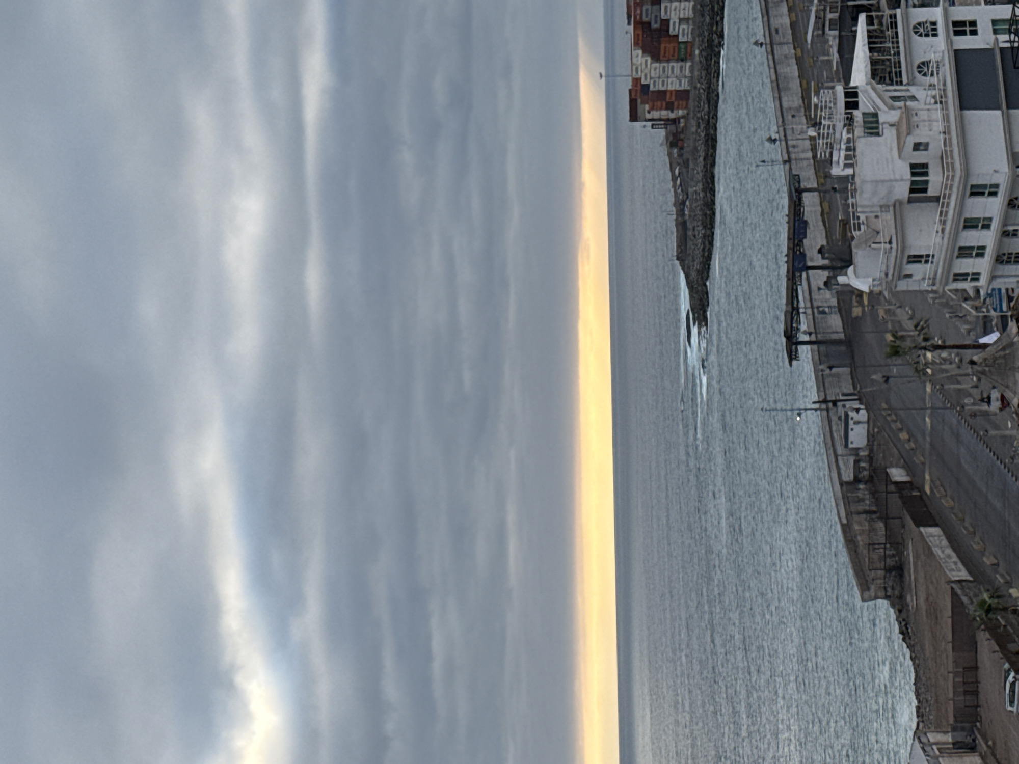 The image depicts a cloudy day scene at the port of Iquique, Chile, overlooking the Pacific Ocean. The sky is filled with gray, overcast clouds, with a faint band of yellow light visible on the horizon where the sky meets the sea, suggesting either sunrise or sunset. The ocean is a deep blue-gray, with visible waves crashing against the rocky shoreline.
In the foreground, the port infrastructure is evident. A multi-story white building, likely an administrative or operational facility, stands prominent on the right side of the image. It features multiple levels, balconies, and windows. Adjacent to this building is a long concrete seawall or quay. Further along the coast, a stack of colorful shipping containers, predominantly white and red, are visible, indicating active cargo operations. A solitary lamppost stands among the containers.
Below the seawall, a paved area with yellow and white markings suggests a roadway or loading zone. A white vehicle is parked in the lower left corner of the image. Vegetation, including palm trees, is sparsely present along the portside. The overall impression is one of a working port under a somber, cloudy sky.