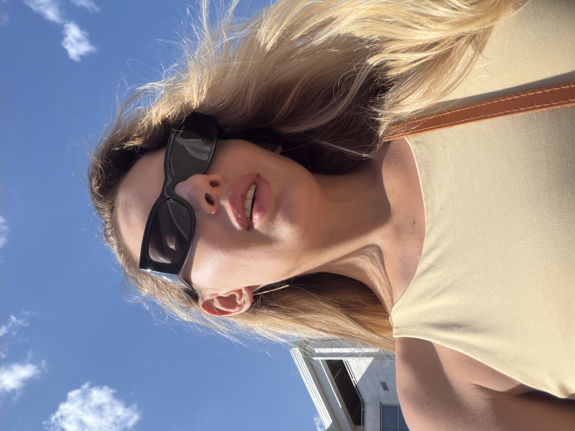 A woman with blonde hair and sunglasses is looking up at the sky. She is wearing a light yellow tank top. Behind her, a building with a light-colored facade and windows is visible. The sky is blue with some white clouds. The scene appears to be outdoors in a sunny location.