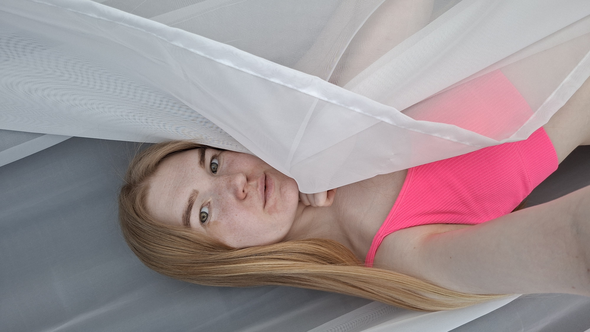 A young woman with fair skin, freckles, and long blonde hair is lying down, partially covered by a white, sheer fabric. She is wearing a bright pink top. The setting appears to be indoors. The photo was taken in the city of Chaikovsky, Russia. The subject is looking directly at the camera with a neutral expression.