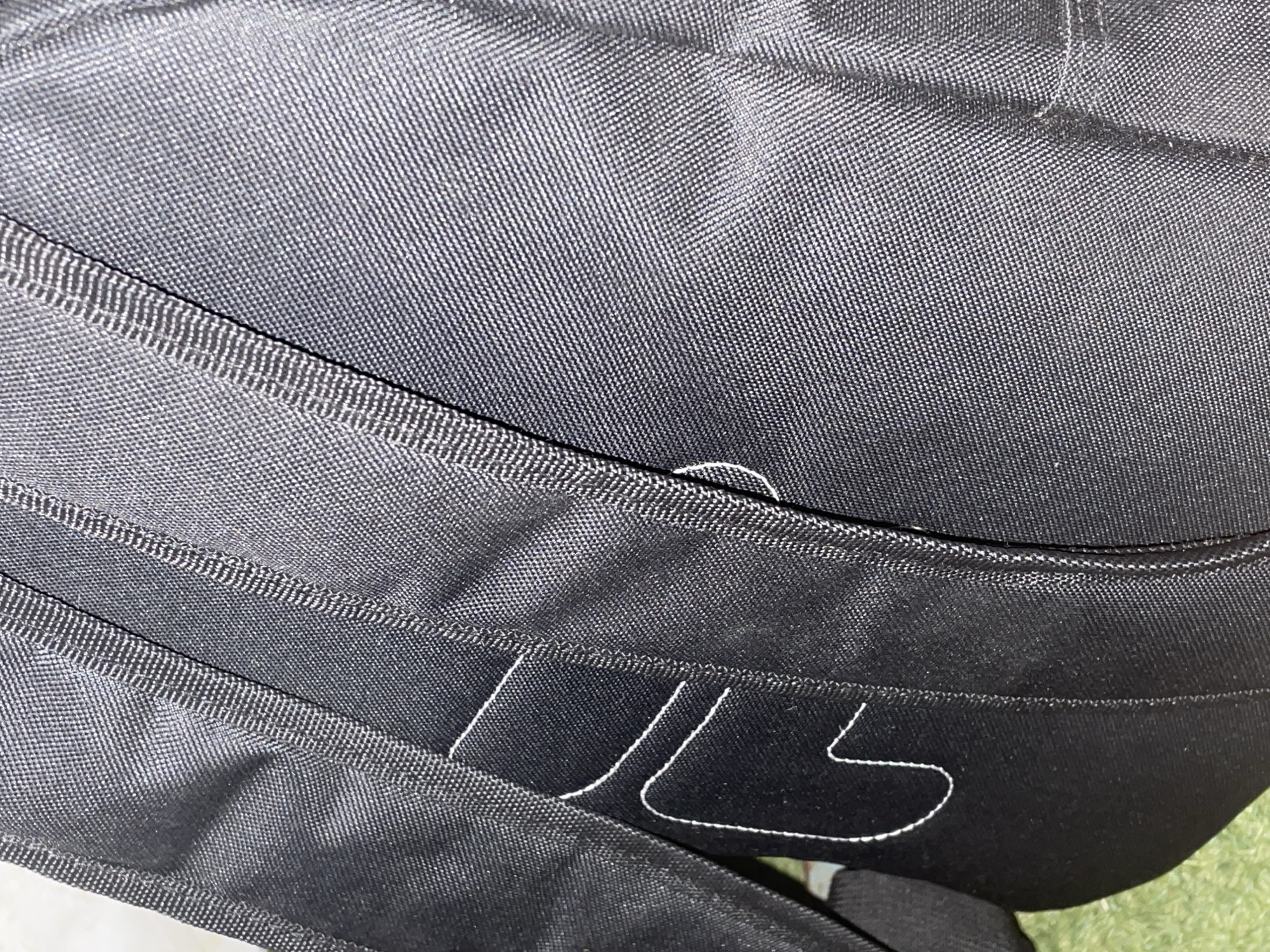 The image presents a close-up, oblique view of a black textile item, identified as a backpack due to visible padded straps and structural seams. The fabric features a uniform, coarse woven texture, consistent with synthetic materials such as nylon or polyester. Prominent black stitching defines edges and panels, with some areas featuring a finer white decorative stitch. A distinct white embroidered logo, resembling a stylized "FILA" design, is partially visible on the left side of the backpack's main body. A wide, black, padded strap is visible along the left edge, curving inwards. The background is out of focus, revealing a blurred green area, likely grass, in the lower left, suggesting an outdoor or partially outdoor environment. The scene is located in the city of Santa Ana, Venezuela.