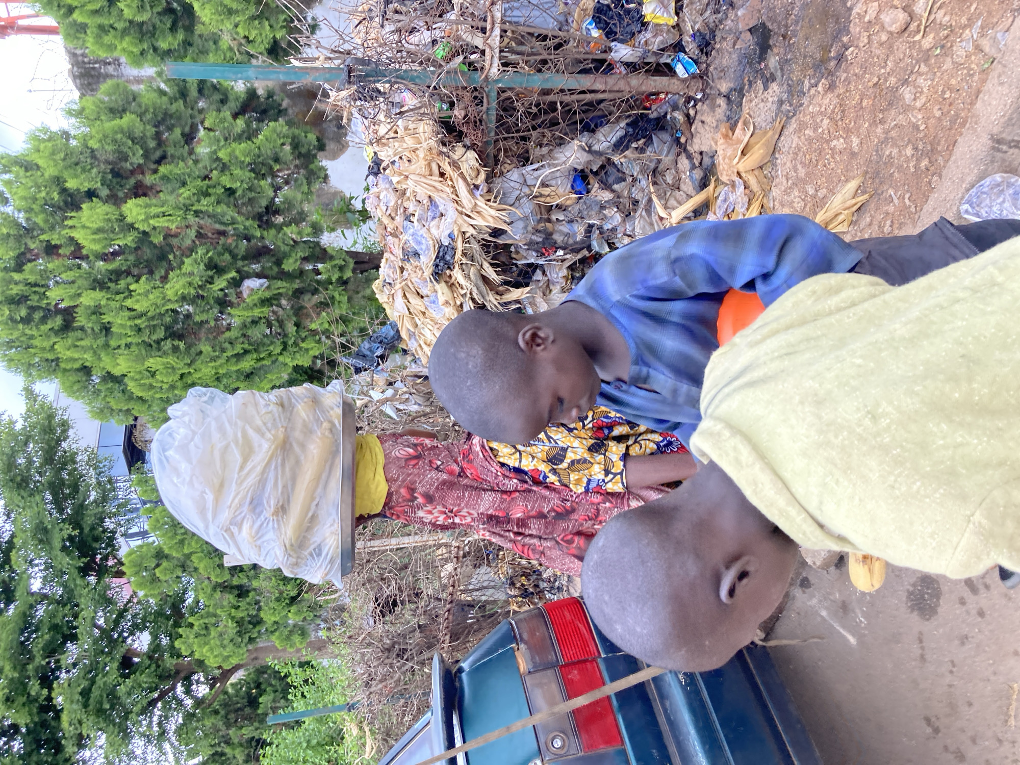 The image shows an outdoor scene in Jos, Nigeria, likely during the daytime with overcast or muted lighting. The setting appears to be a somewhat unkempt area next to a road, with a large pile of garbage and debris visible. In the foreground, two young boys are the primary subjects. One boy, wearing a blue and black plaid shirt, is looking down and appears to be holding an orange object. He is positioned to the right of the image. Another boy, whose head and shoulder are visible in the lower right, is wearing a light green garment.
Behind the boys, a woman is visible, partially obscured by a large, clear plastic bag that she is carrying on her head. She is wearing a colorful patterned dress with a red and white floral print and a yellow headscarf. A metal tray or base is beneath the plastic bag.
The background is dominated by a dense green tree on the left and the pile of refuse on the right, which includes various types of plastic bags, dried plant matter, and other refuse. A metal fence or barrier separates the area with the garbage. Part of a dark green vehicle, specifically its taillight assembly, is visible in the lower part of the image. The overall impression is of a busy street scene in an urban or semi-urban environment, where waste disposal may be a challenge. No text is visible in the image. The expressions on the faces of the children are not clearly discernible, but their posture suggests they are engaged in their activities.