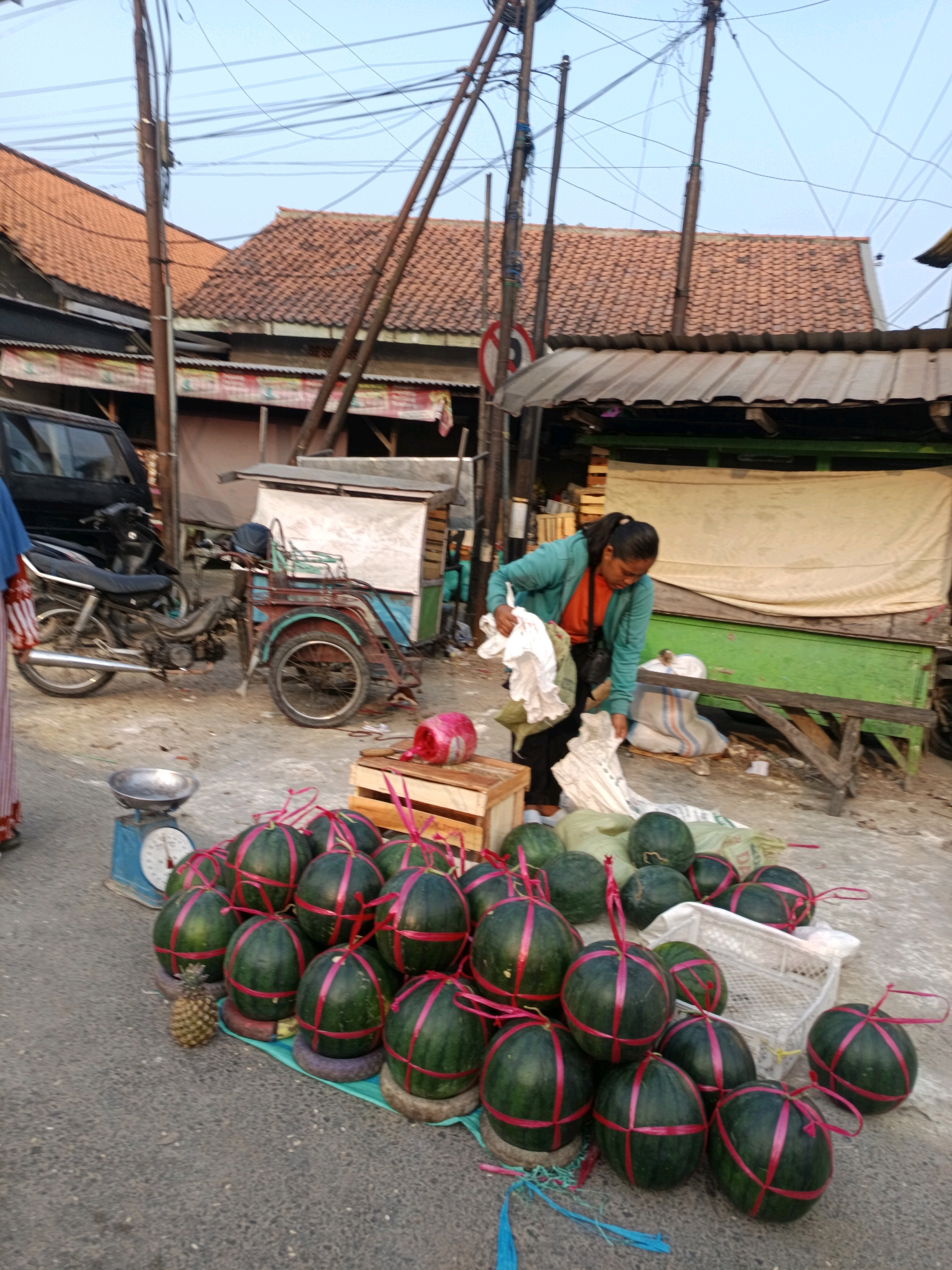 In a street market in Blega, Indonesia, a woman in a teal hooded jacket and orange shirt is seen arranging produce. She is standing next to a collection of watermelons, some of which are tied with pink ribbons. A small pineapple is also visible among the watermelons. In the background, a motorcycle and a three-wheeled cart are parked. Buildings with tiled roofs line the street, and power lines crisscross overhead. The scene suggests a typical day at an outdoor market.